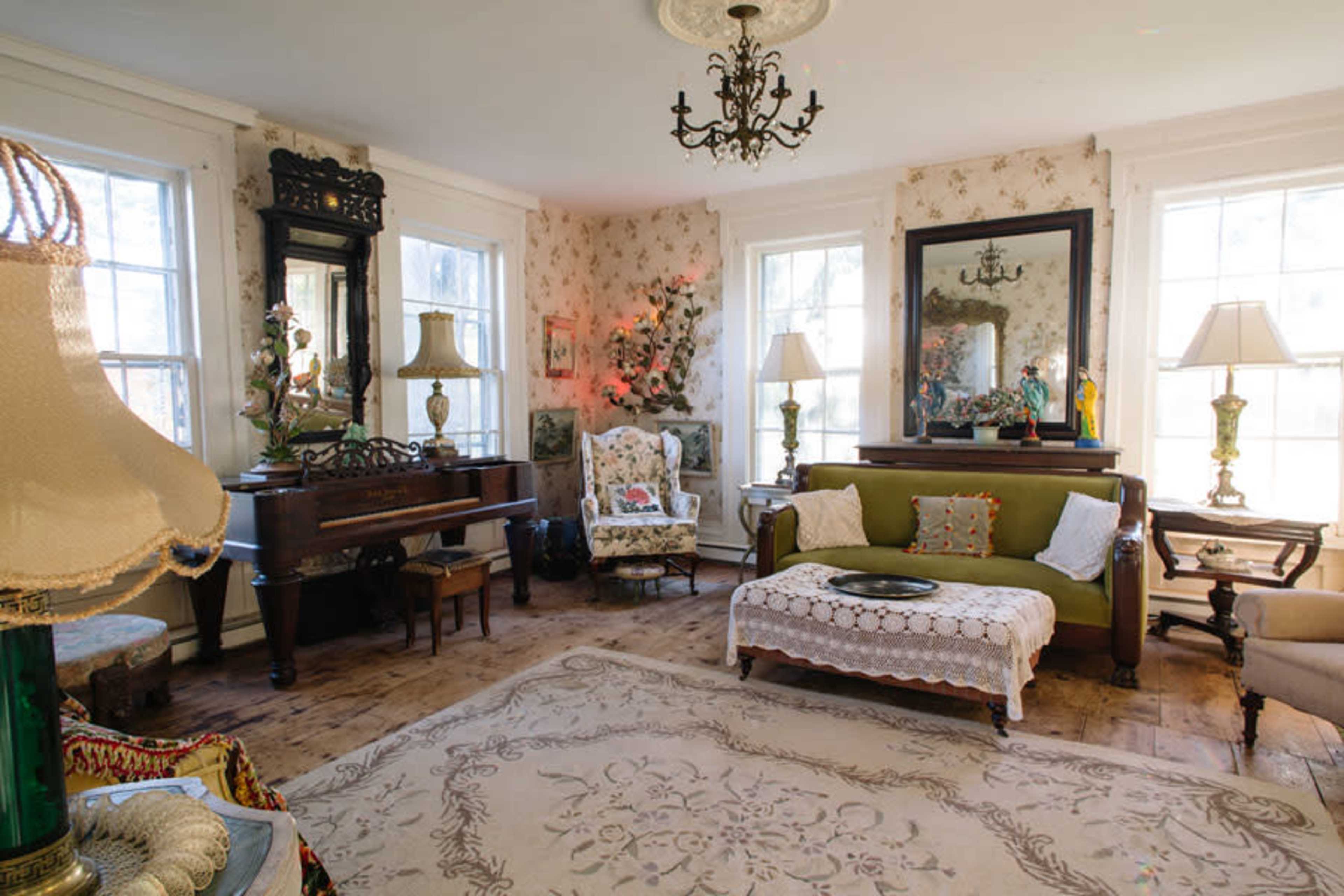 The room features a mix of vintage furniture, including a green sofa, a wooden piano, and decorative lamps, surrounded by patterned walls and large windows.