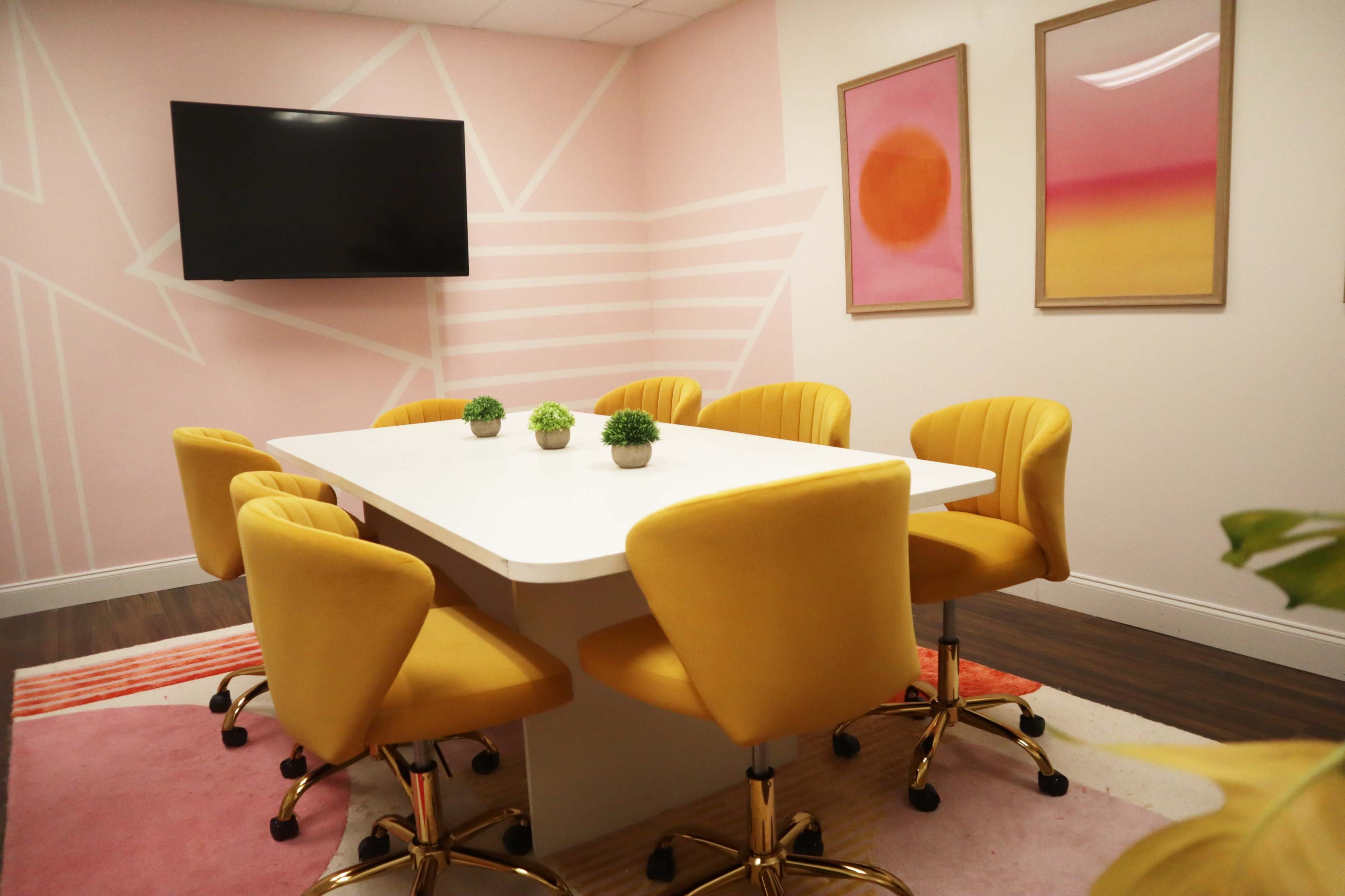 A modern conference room with a white table surrounded by eight yellow swivel chairs, a large wall-mounted TV, and colorful artwork.