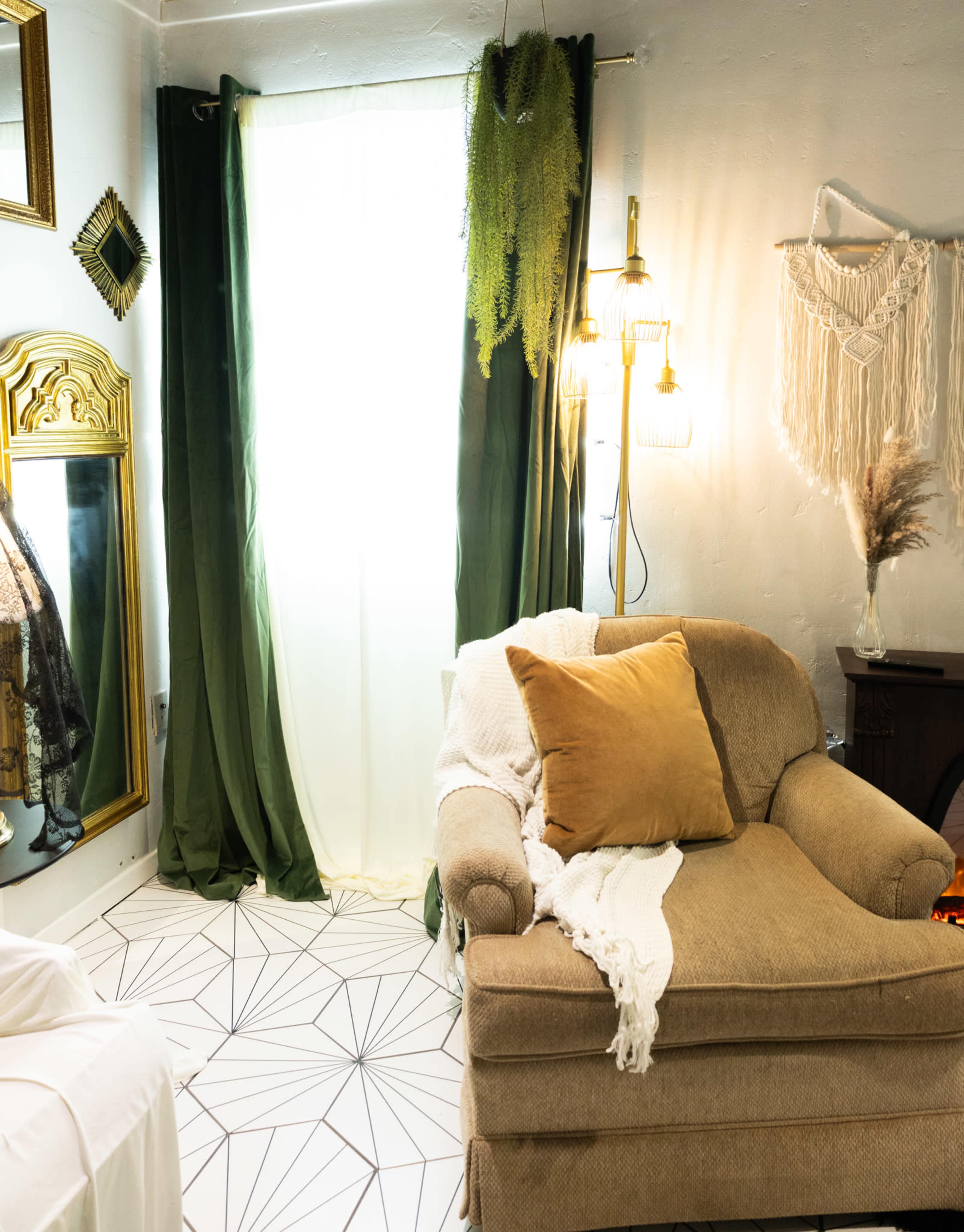 The image shows a cozy interior space featuring a beige armchair with a brown cushion, a white throw, and green curtains, alongside a floor with geometric patterns and a large mirror reflecting the light.