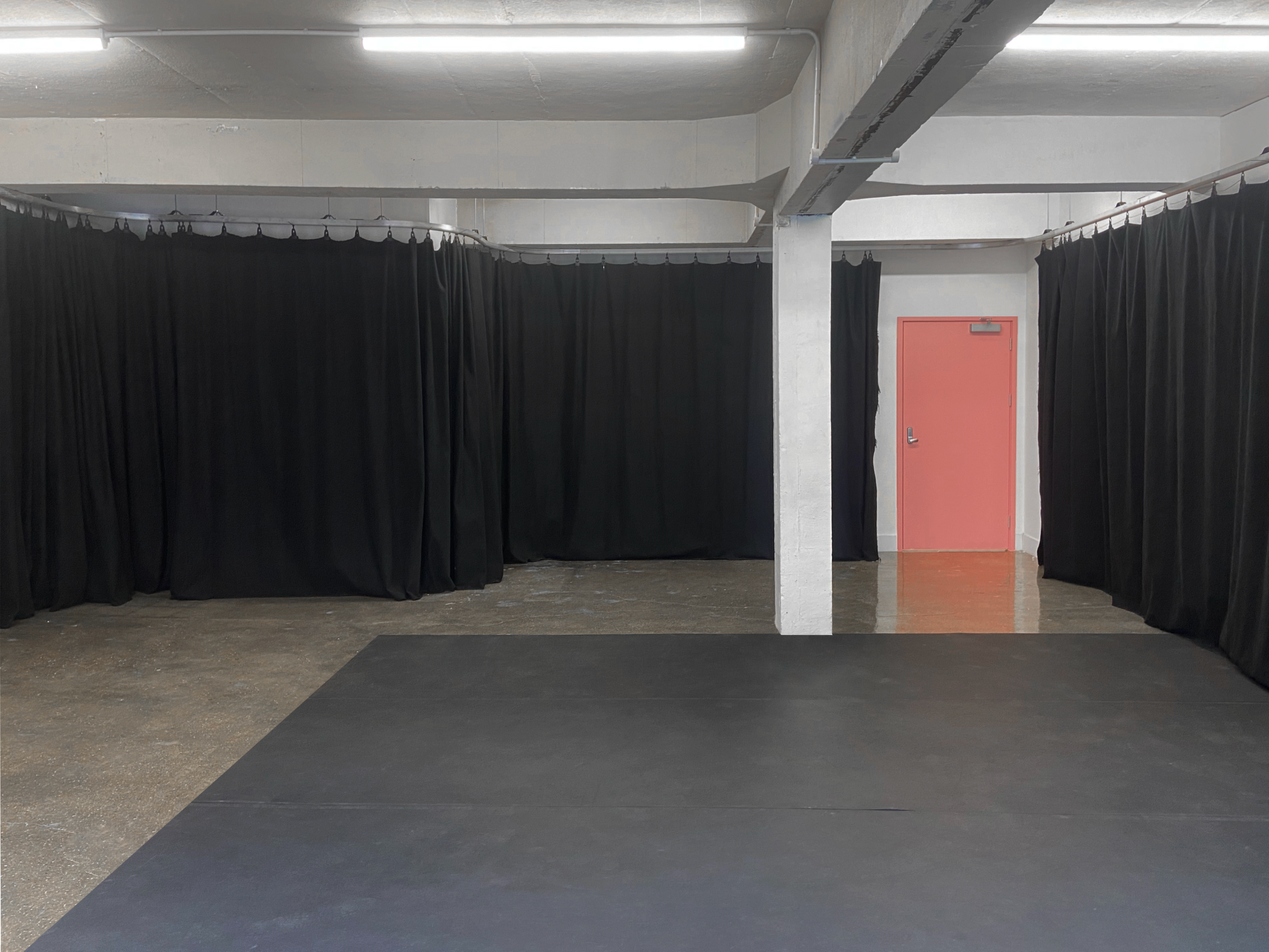 A spacious room with black curtains lining the walls, a pink door at the far end, and a dark floor surface.