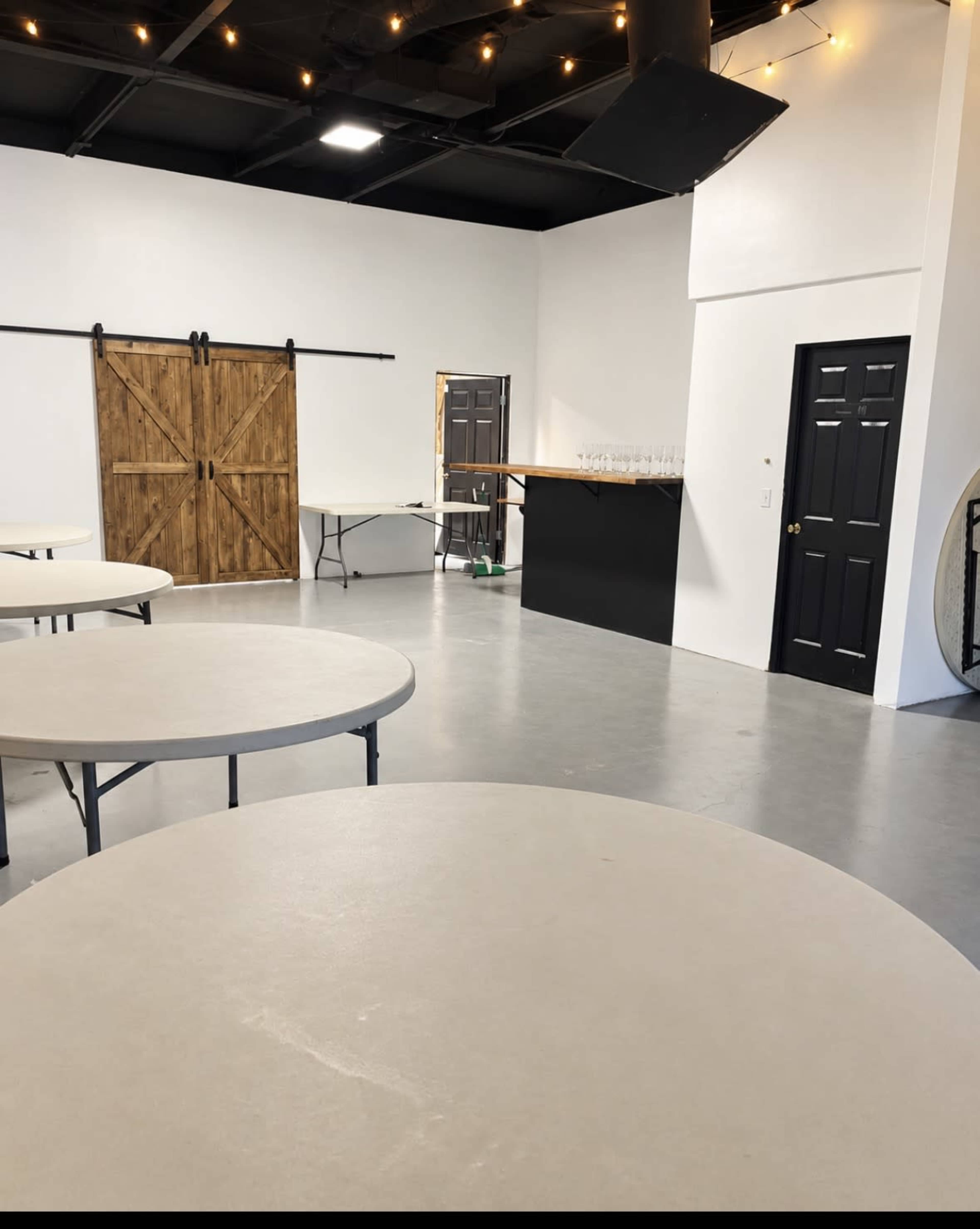 The image shows an empty event space with several round tables, a bar area in the background, and a wooden sliding door.