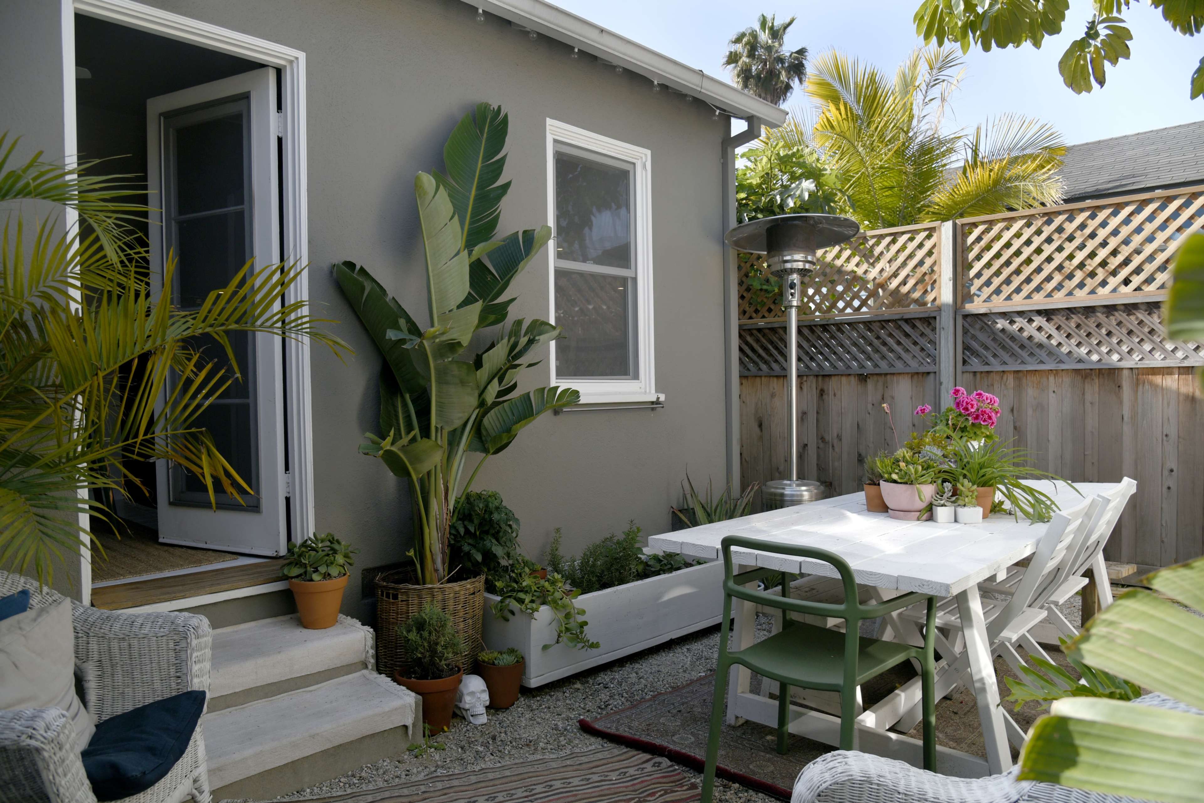 A patio features a white dining table surrounded by chairs, potted plants, and a doorway leading into a house, with a heat lamp and palm trees in the background.