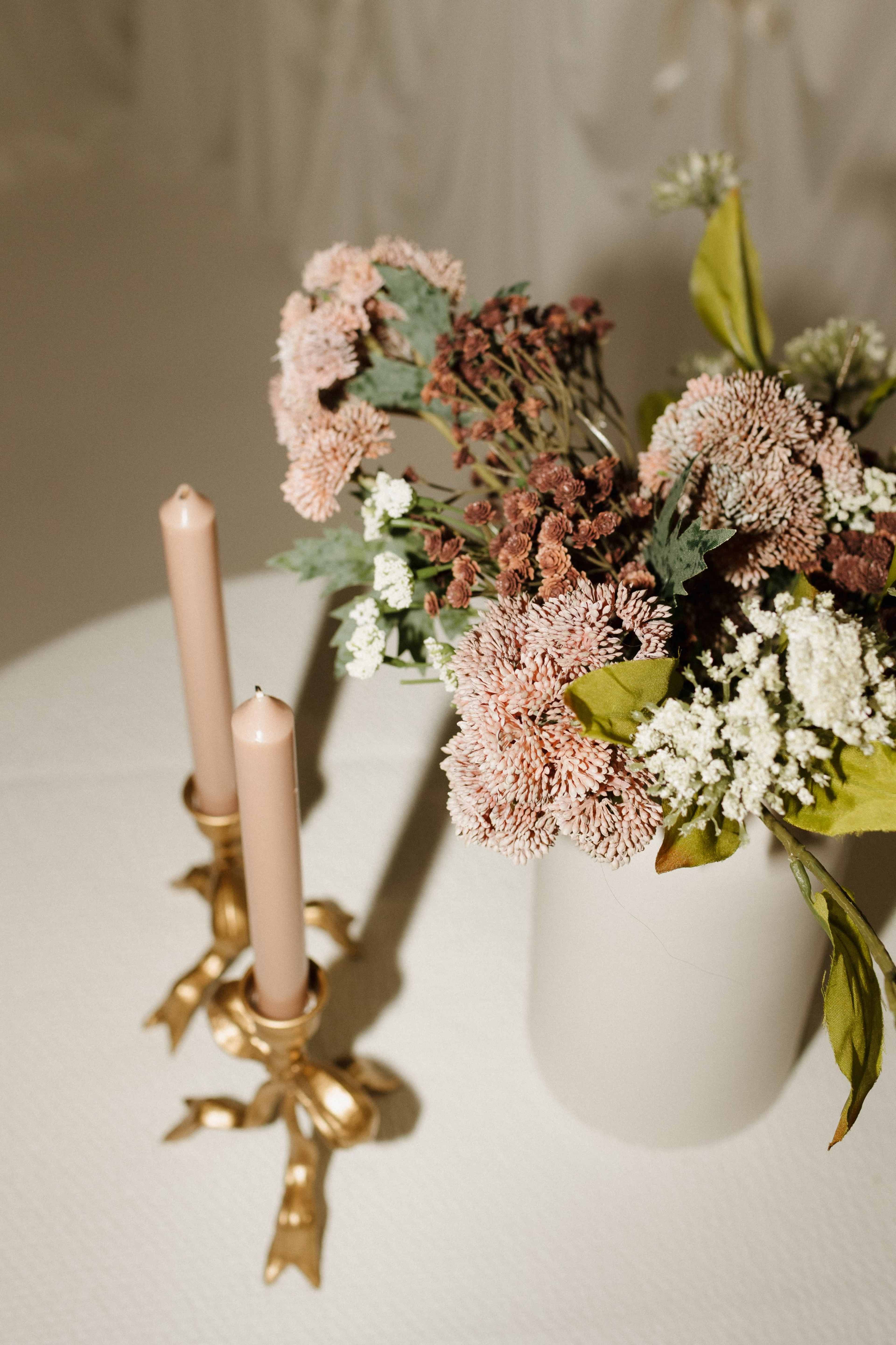 A vase filled with dried flowers stands beside two pink candles in a gold candle holder on a textured surface.