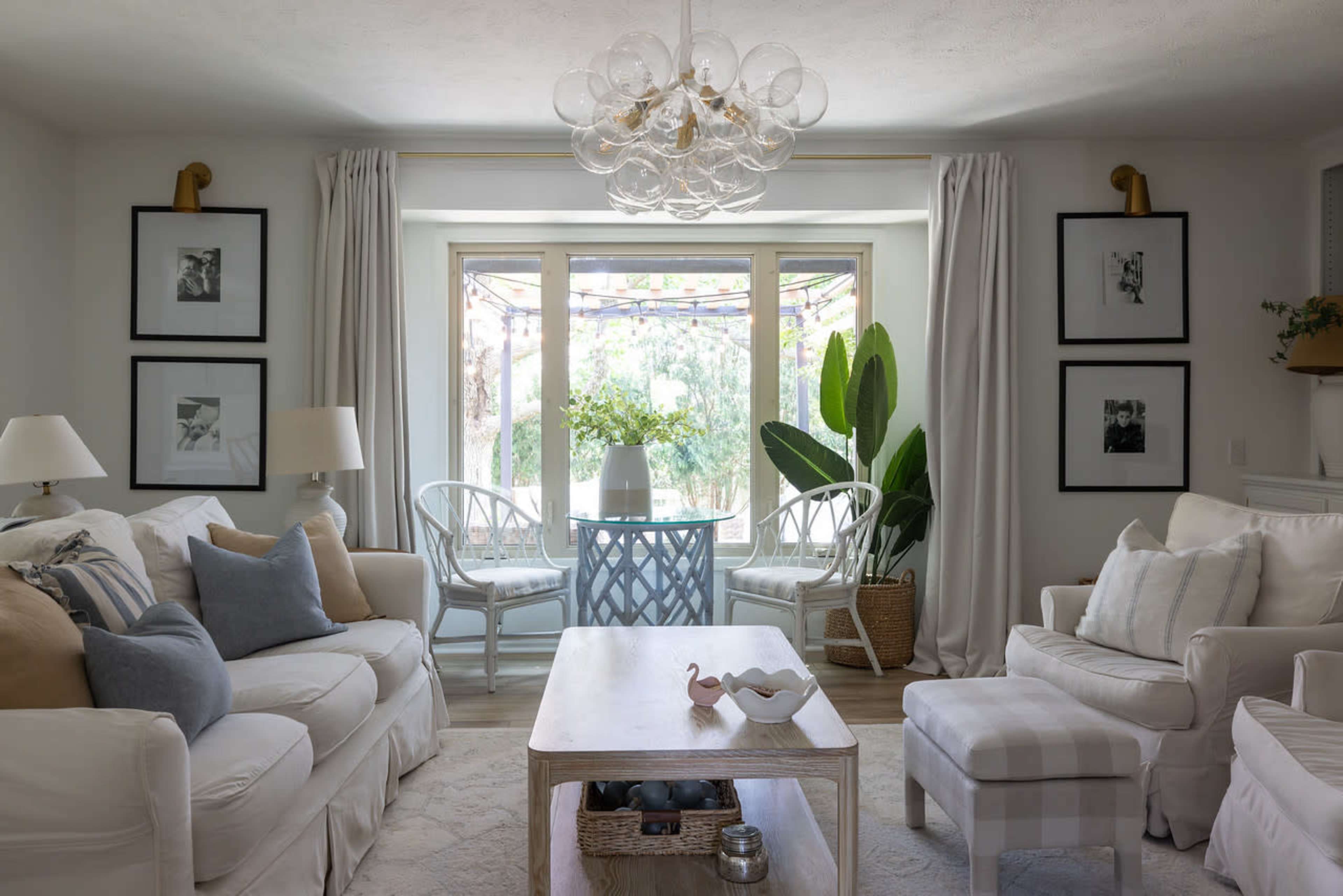 The image shows a cozy living room with white sofas, framed black-and-white photographs on the walls, and large windows adorned with sheer curtains overlooking greenery.