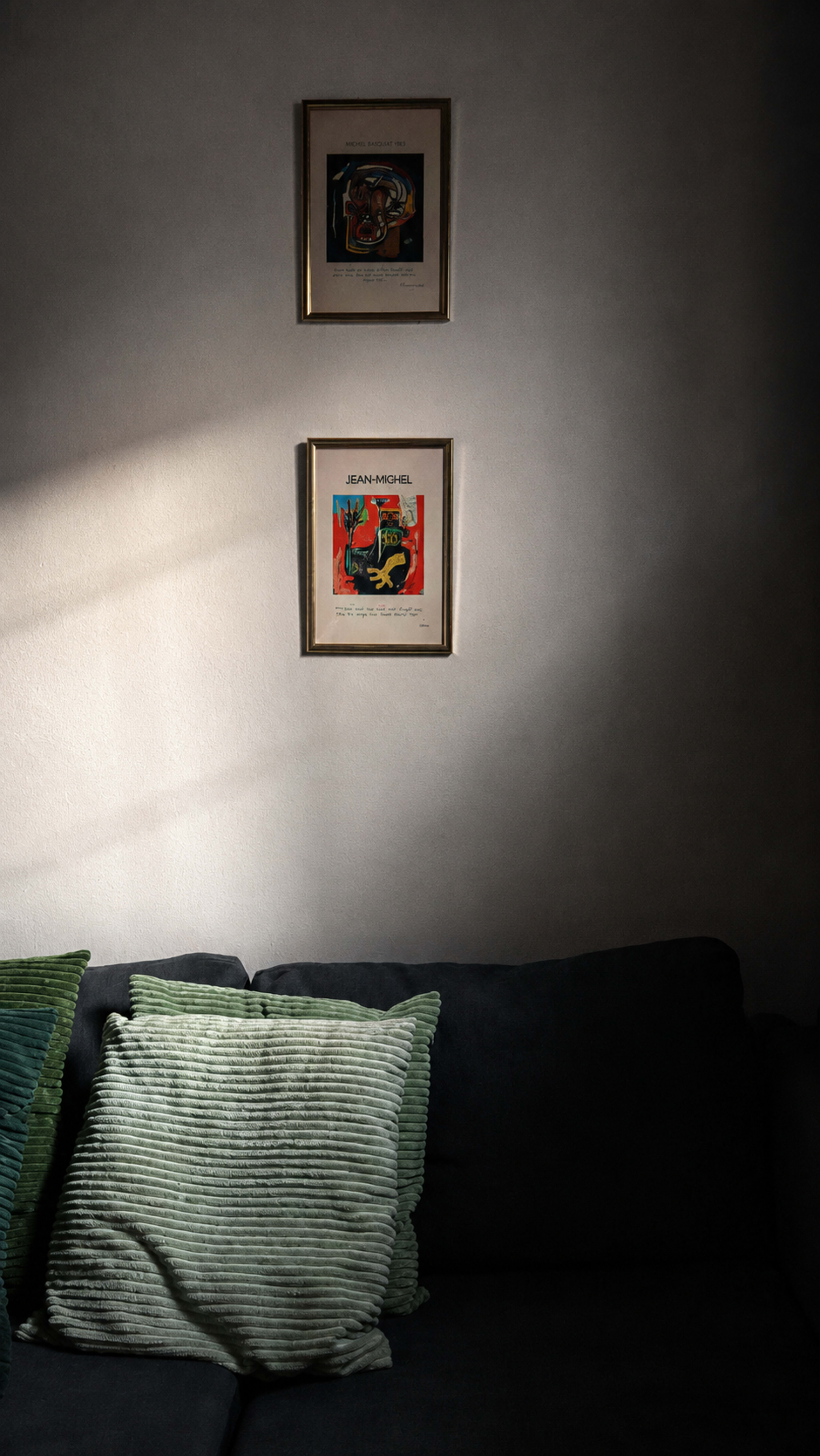 The image shows a darkly lit living room corner featuring a grey couch with green textured pillows and two framed artworks hanging above it.