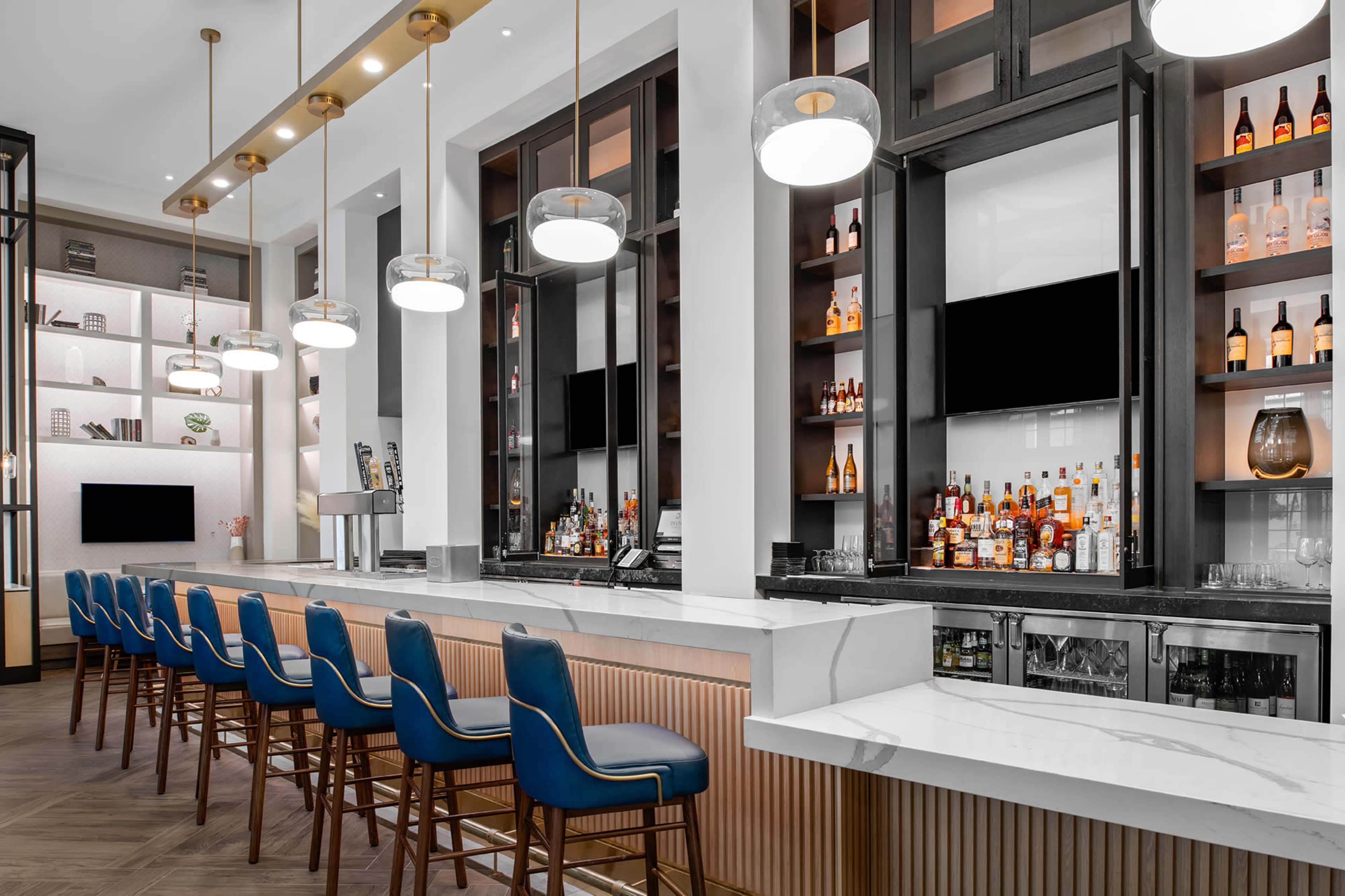 The image shows a modern bar area featuring a long marble counter, high-backed blue chairs, and shelves with various bottles and decor.