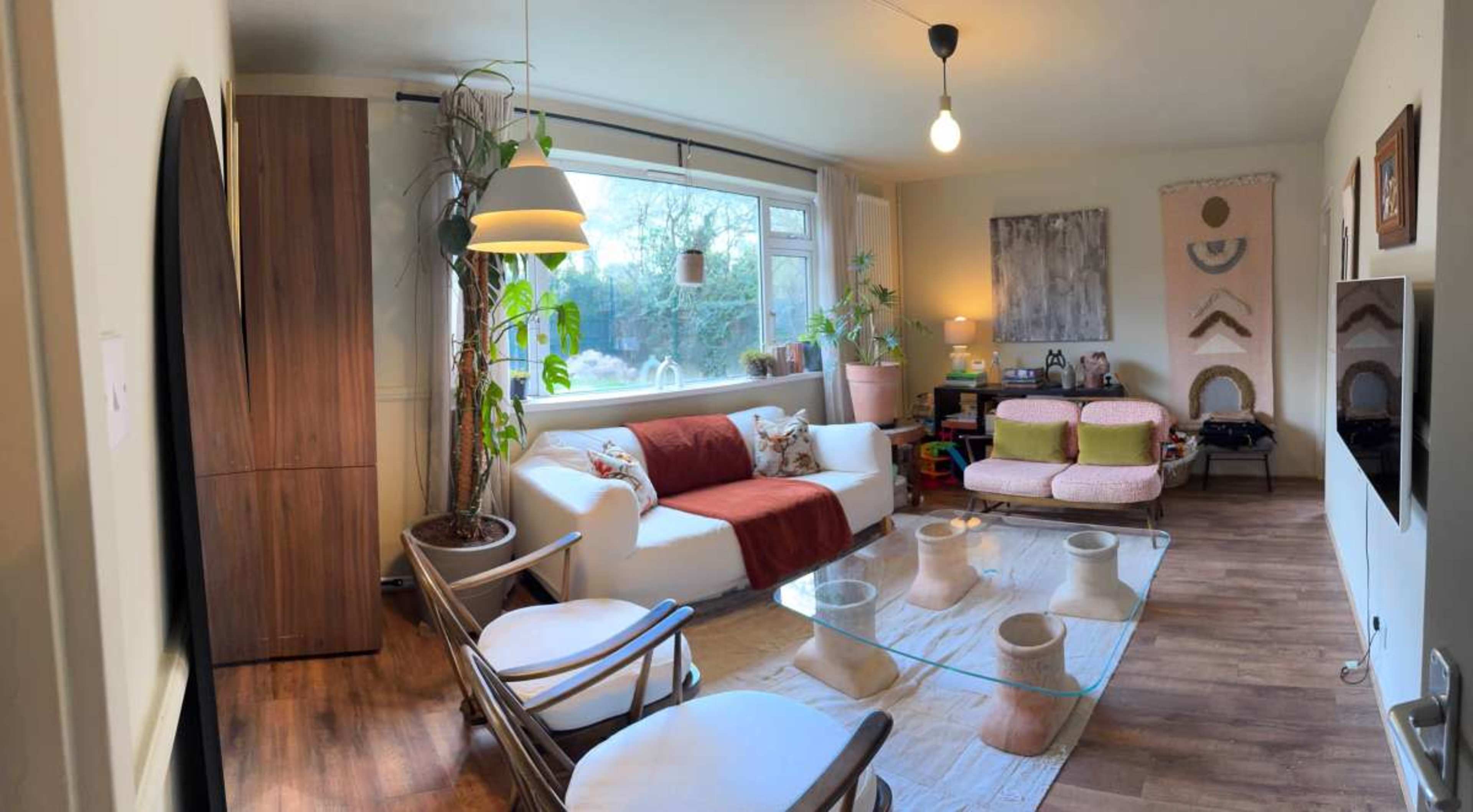The living room features a white sofa, a glass coffee table, and various plants, with large windows letting in natural light.