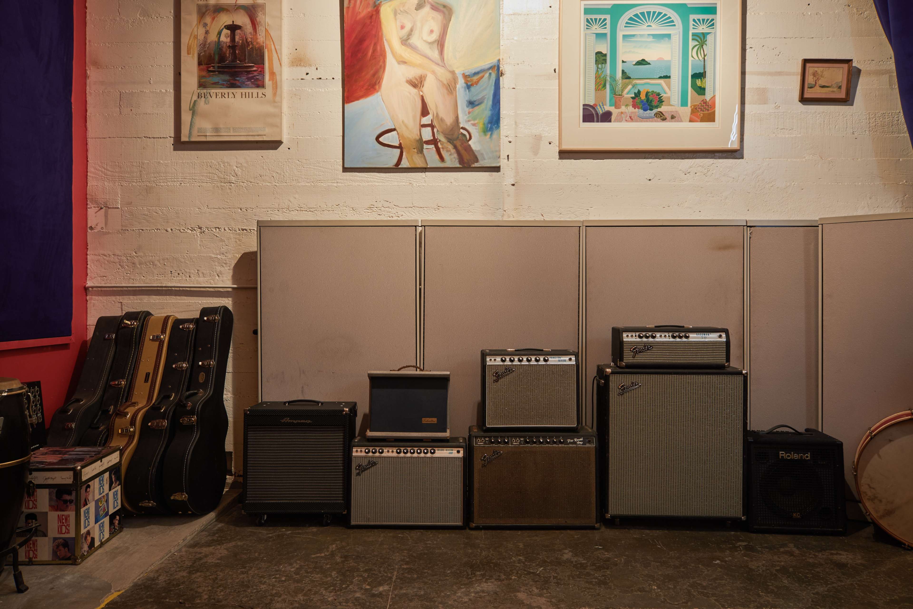 The image shows a wall with various amplifiers stacked on the floor beside several guitars leaning against a partition, with framed artwork hanging above.