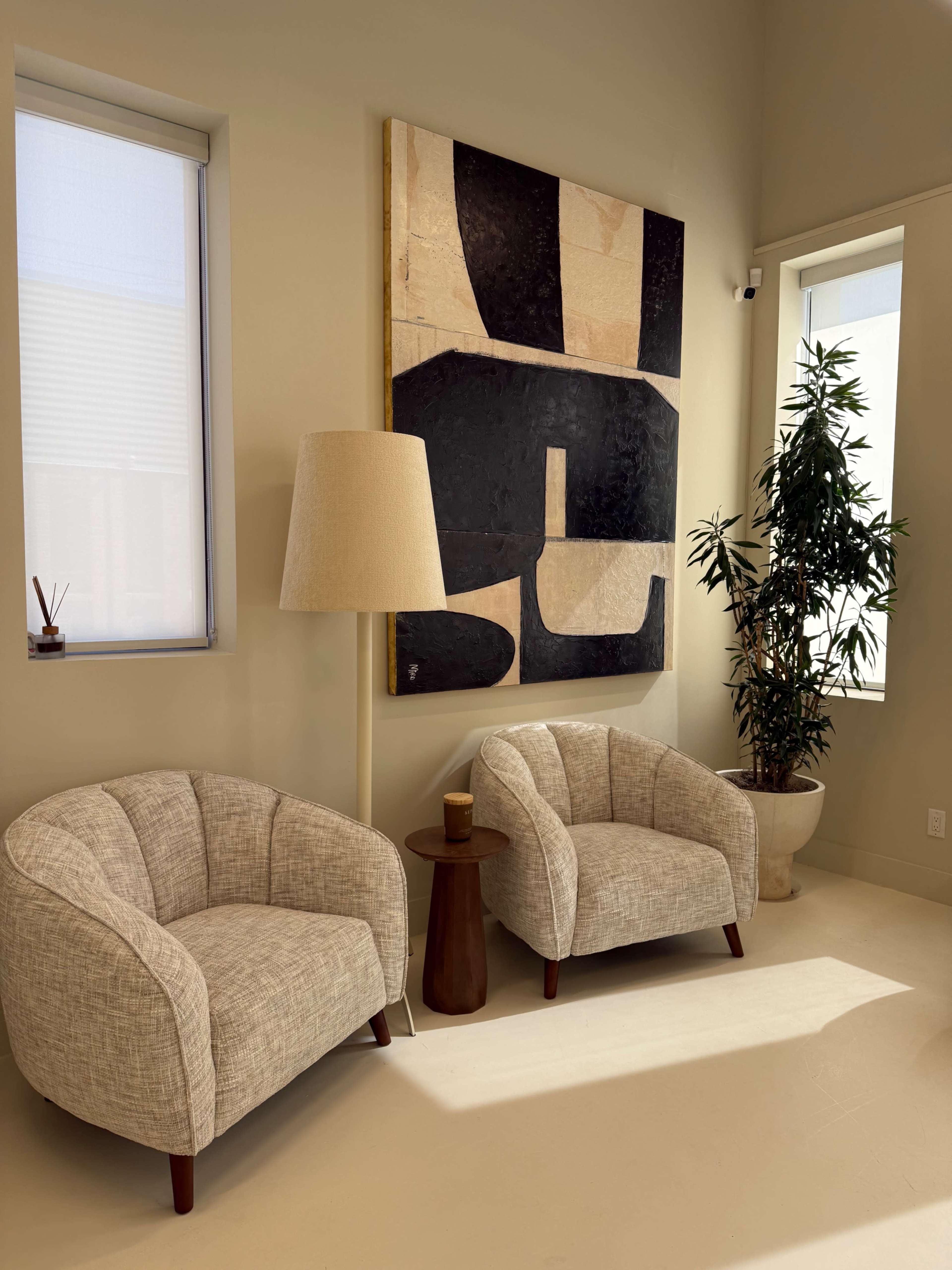 The image shows a bright, modern interior featuring two comfortable beige armchairs, a small side table, a tall lamp, and a large abstract painting on the wall, alongside a potted plant.