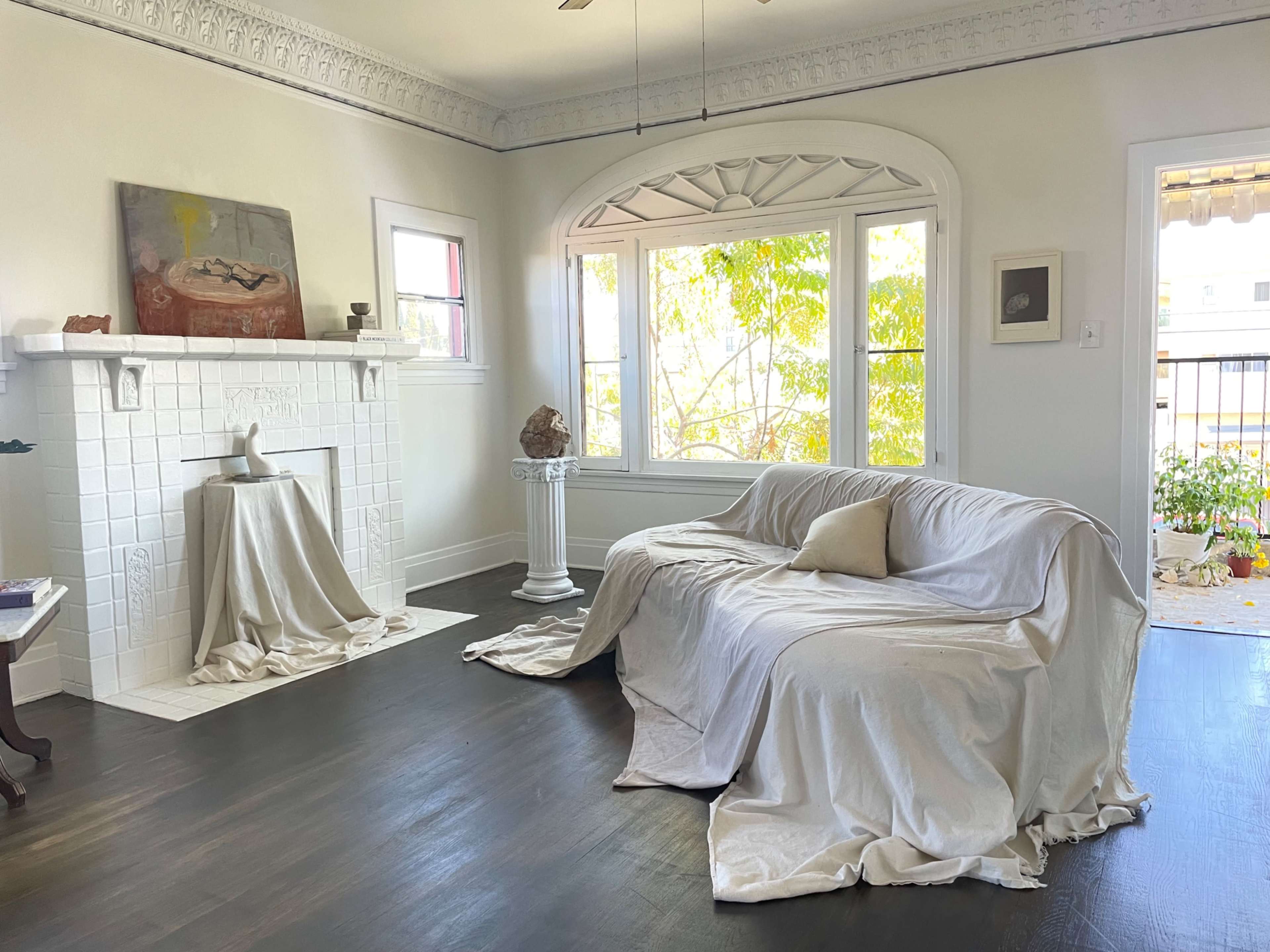 The living room features a white-covered couch, a sculptured pedestal beside it, and large windows letting in sunlight, illuminating the space.