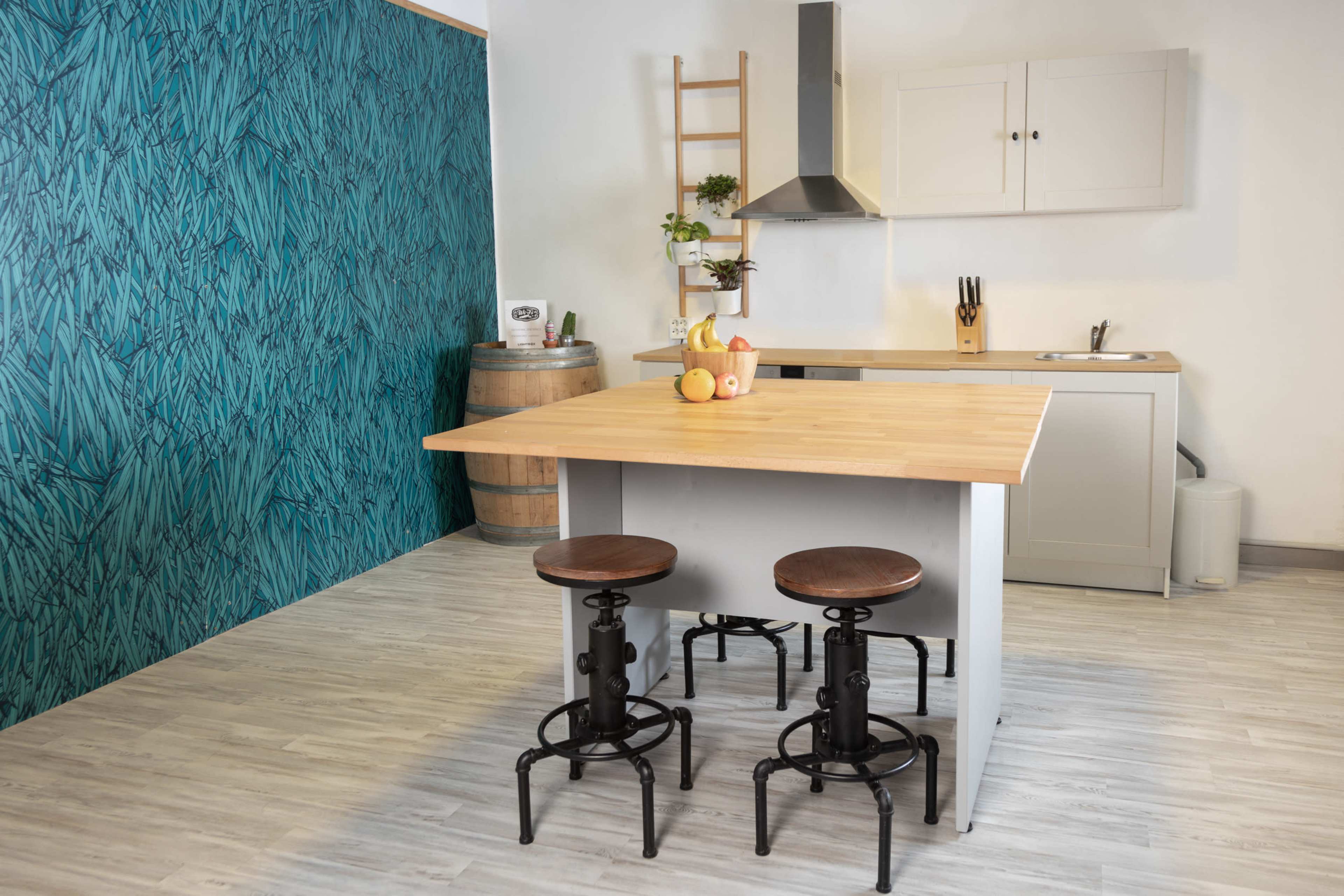 A modern kitchen features a wooden table with two metal stools, light cabinetry, and a green patterned wall.