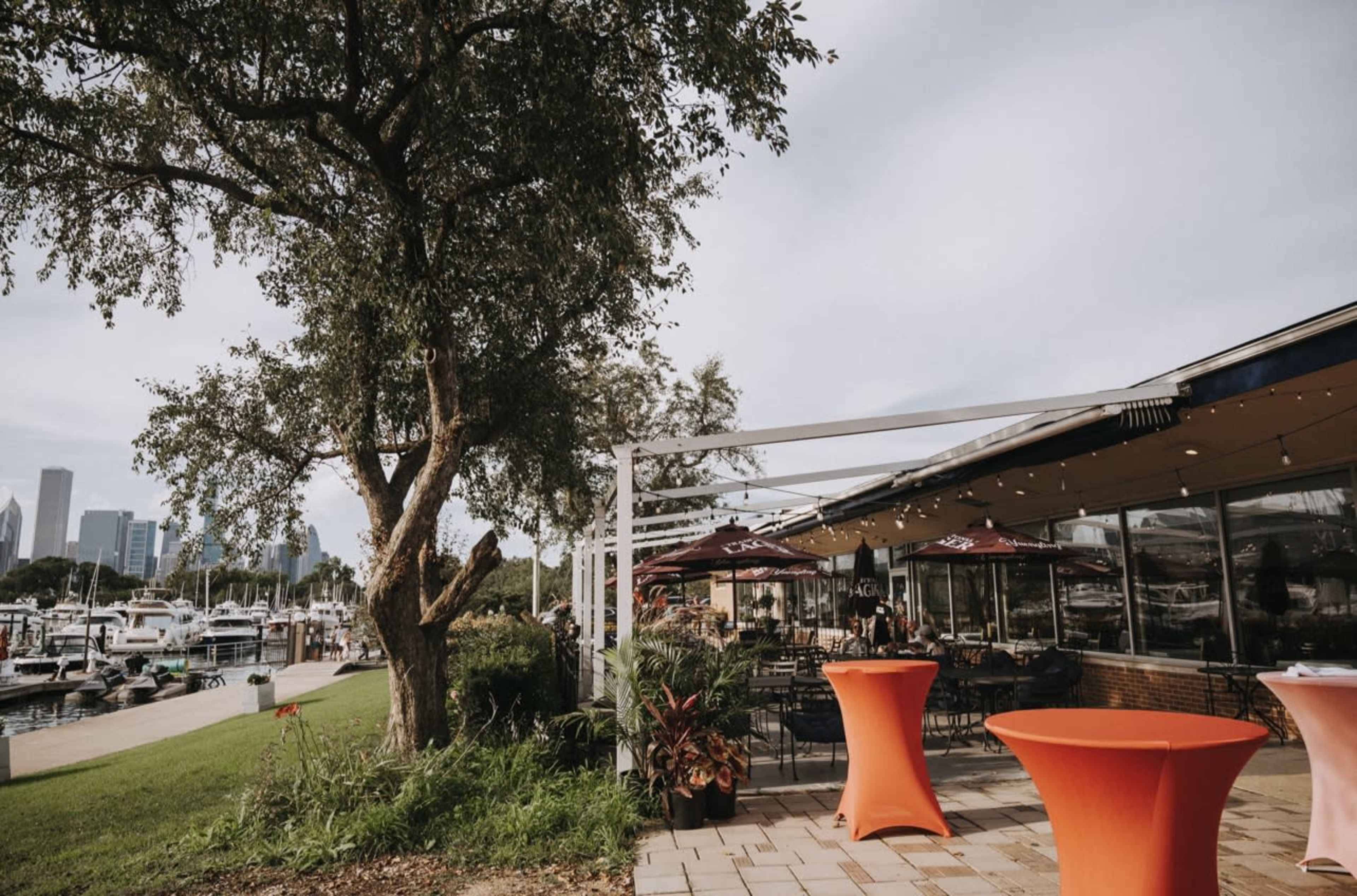 The image shows an outdoor dining area with orange tables, a large tree, and a marina with boats in the background against a city skyline.