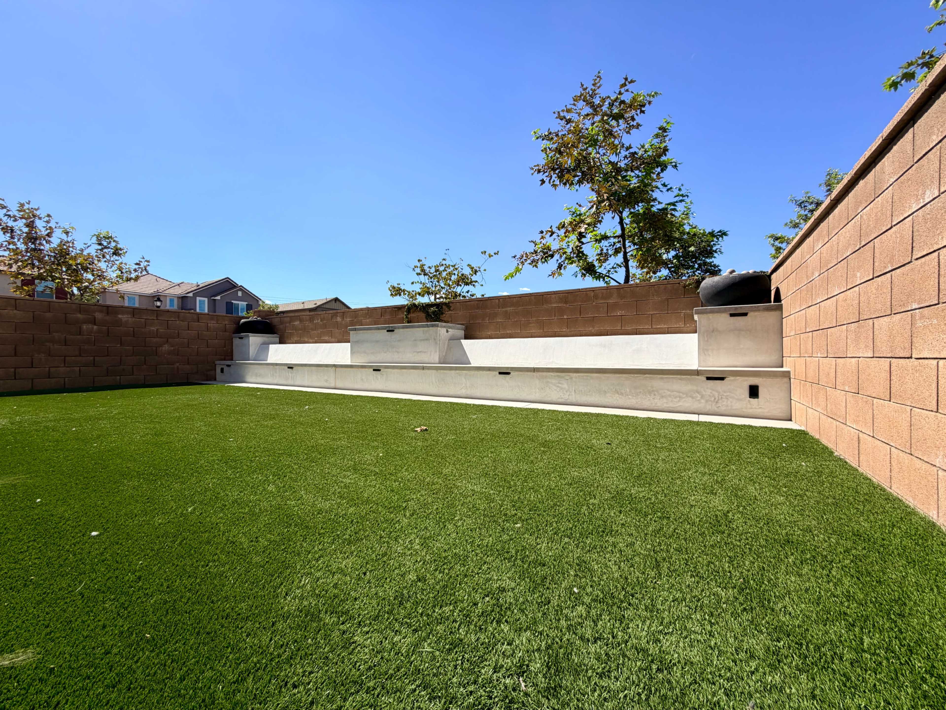 The image shows a landscaped outdoor area with artificial grass, a concrete seating structure along a wall, and trees in the background.