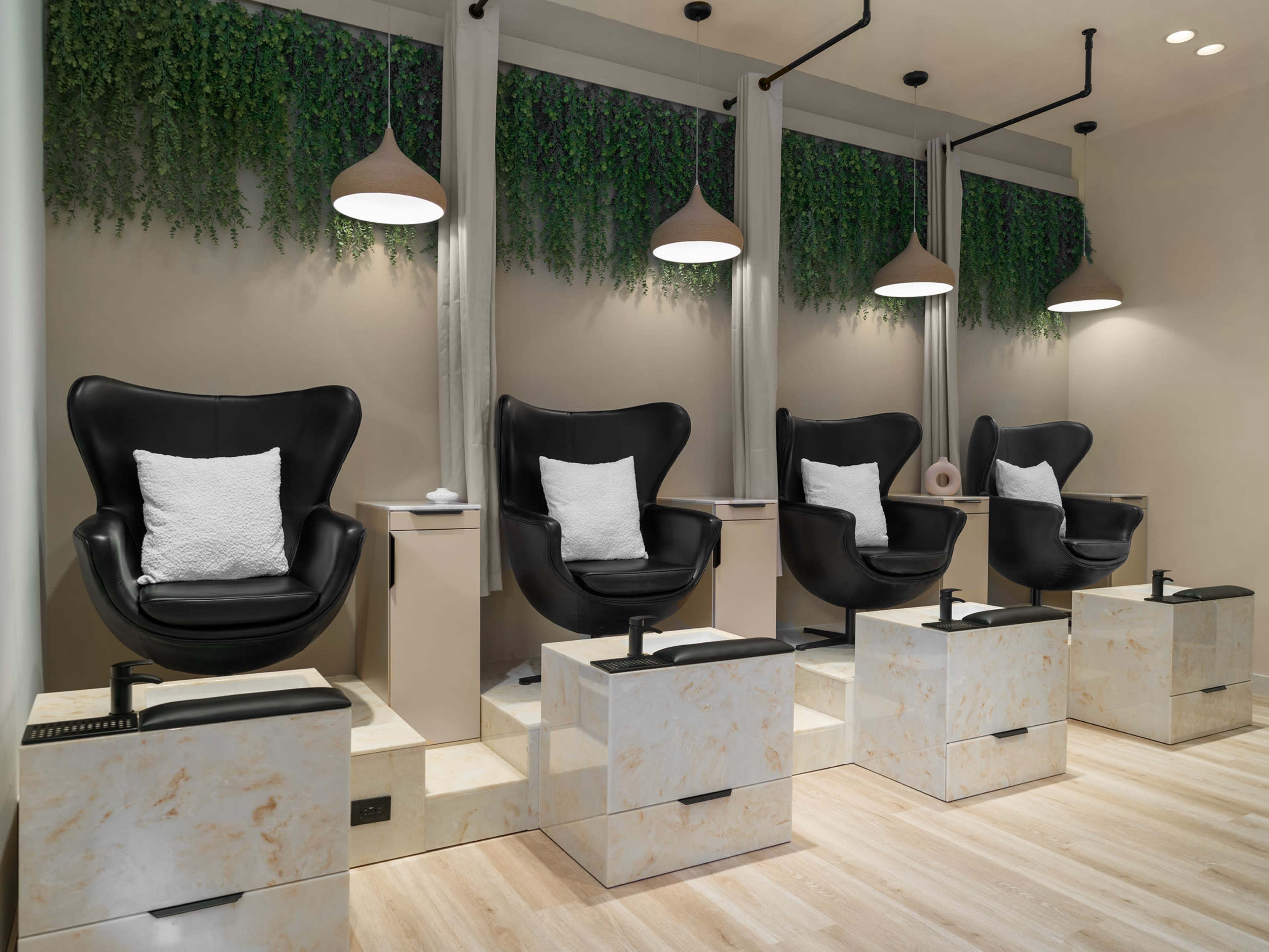 A row of elegant black pedicure chairs with white cushions is arranged against a light-colored wall, complemented by hanging pendant lights and greenery.