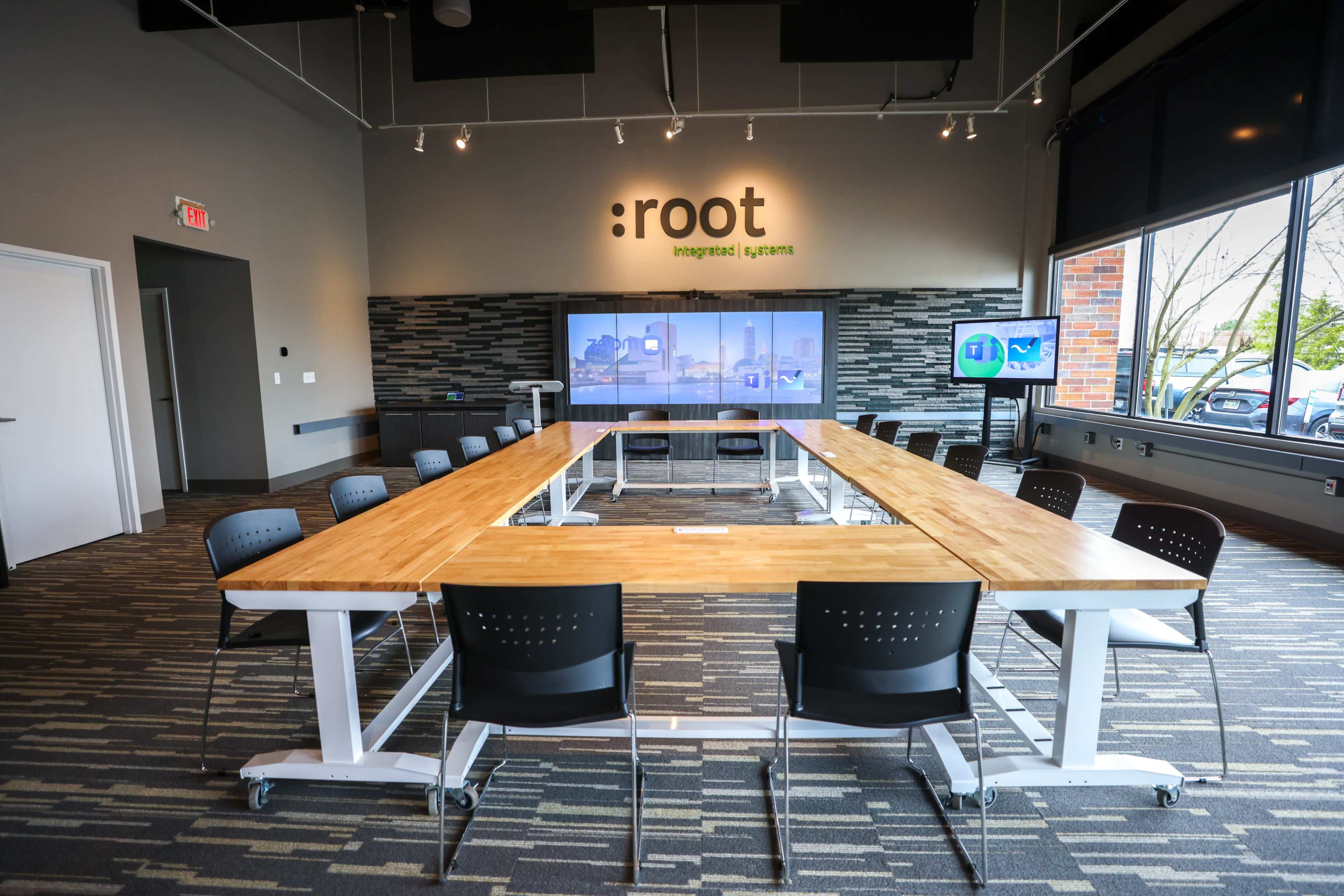 A conference room features a long wooden table surrounded by black chairs, with a screen displaying information on one wall.