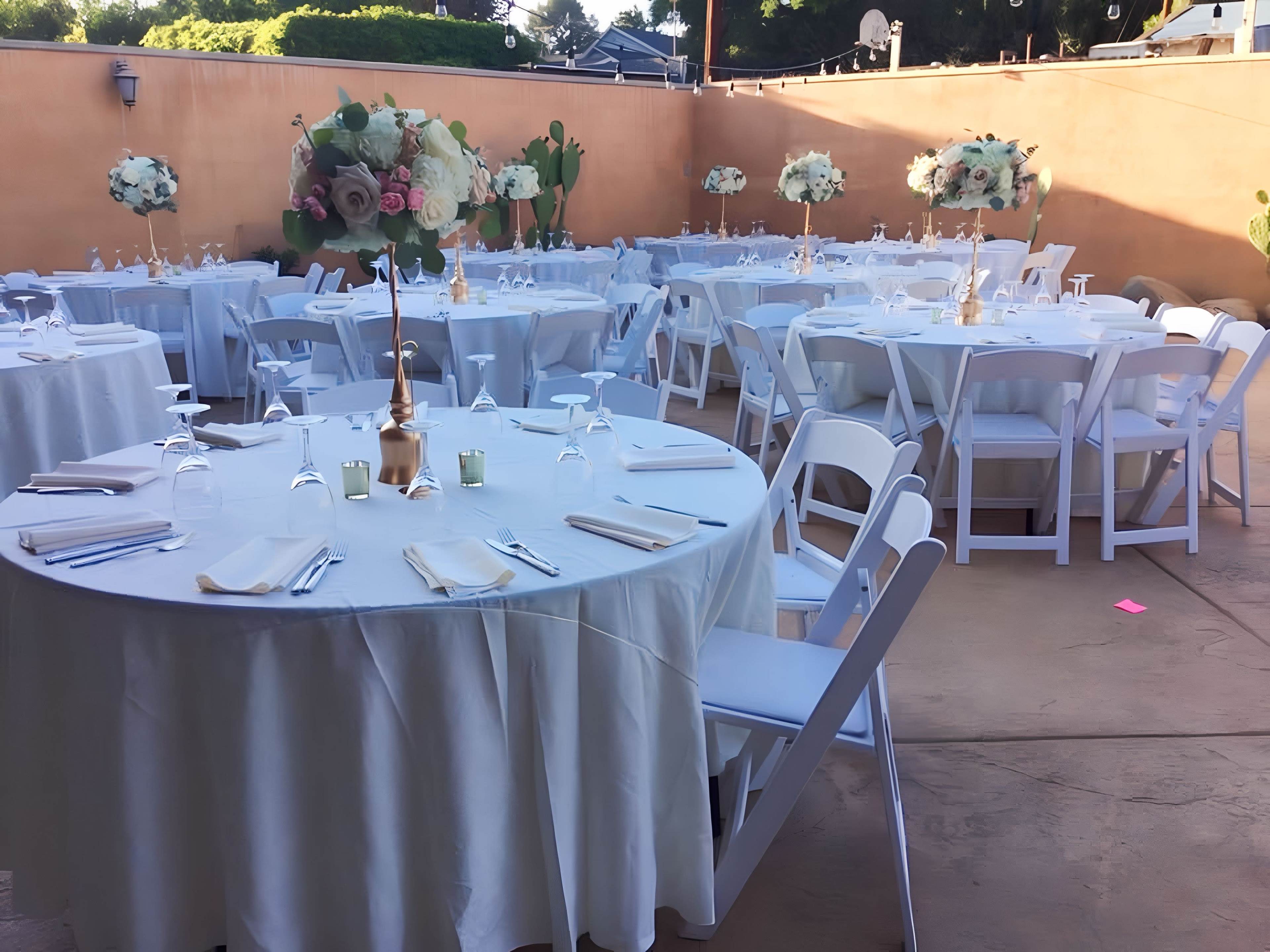 The scene shows a spacious outdoor venue set up for an event, featuring round tables covered with white tablecloths and decorated with floral arrangements.