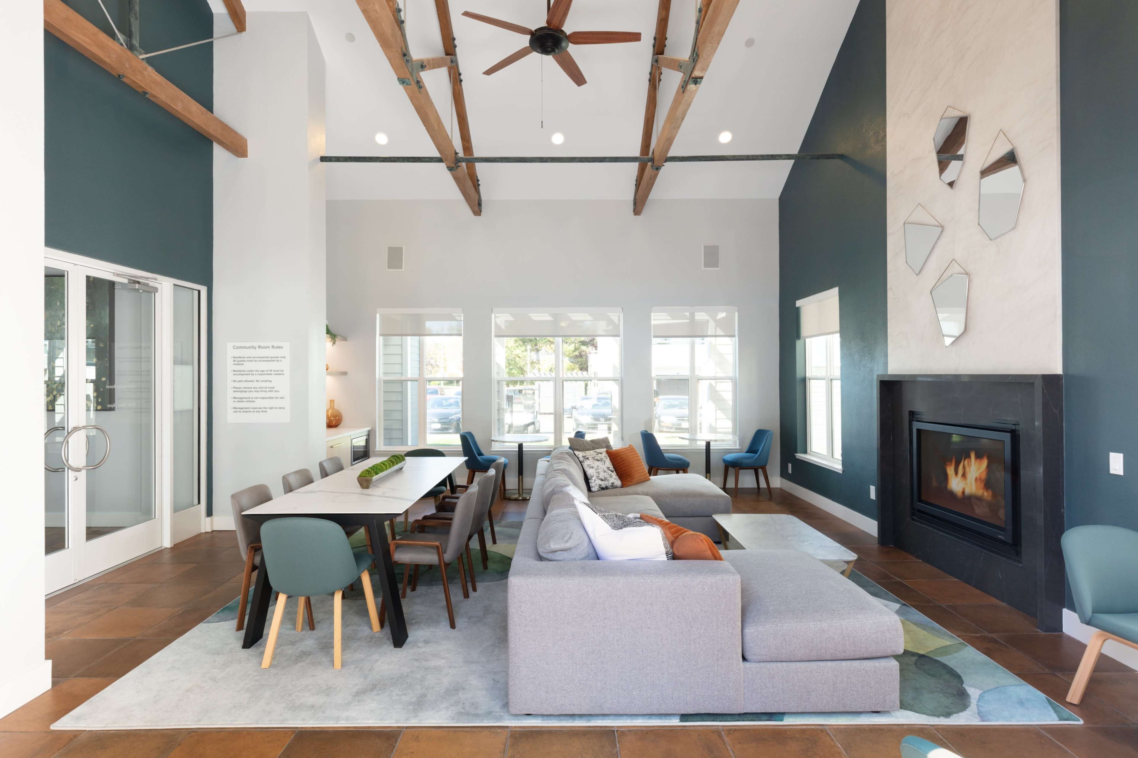 The image shows a modern lounge area with a sectional sofa, dining table, and a fireplace, featuring high ceilings with exposed wooden beams and large windows.