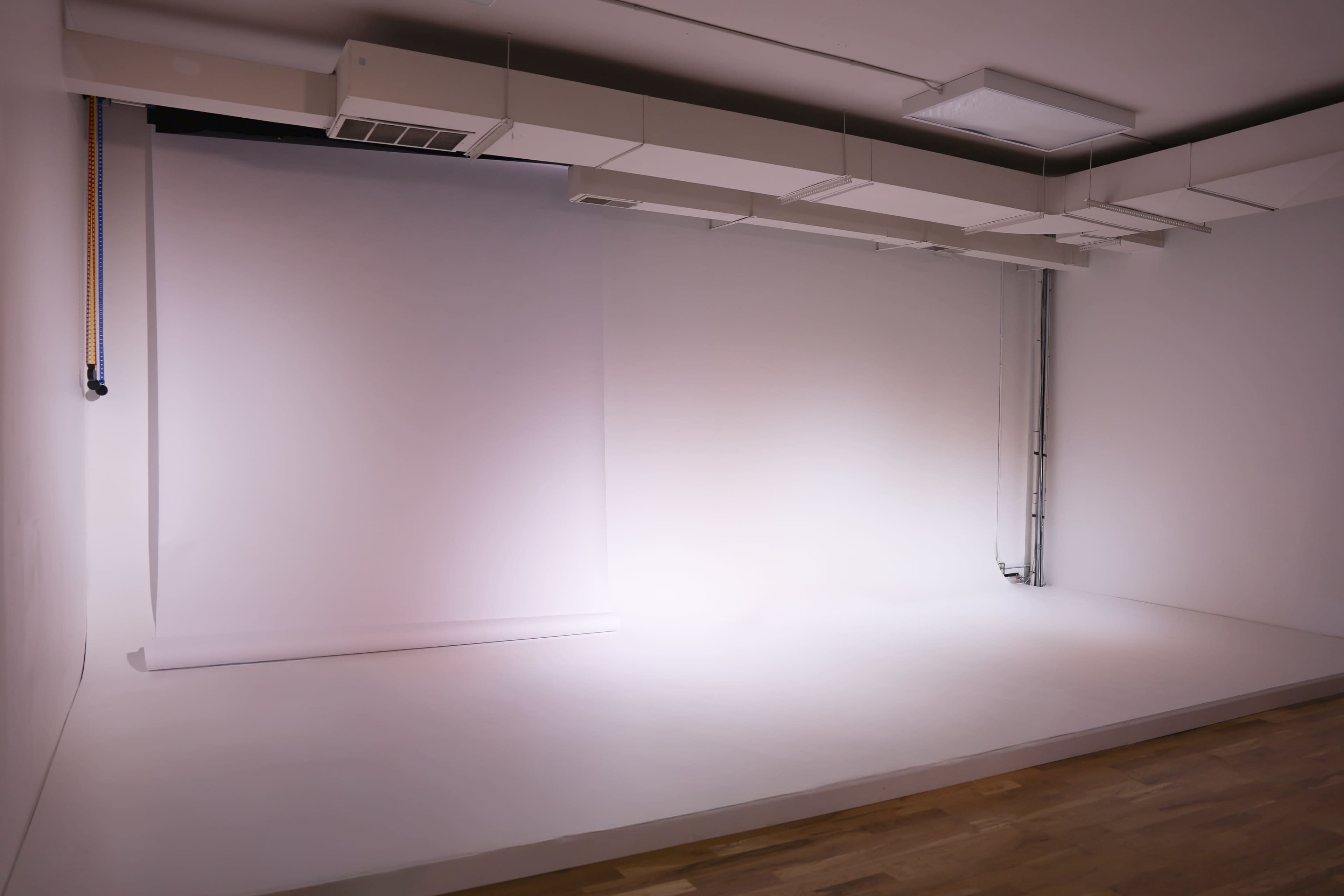 The image shows a minimalist photography studio with a blank backdrop, lighting fixtures, and a wooden floor.