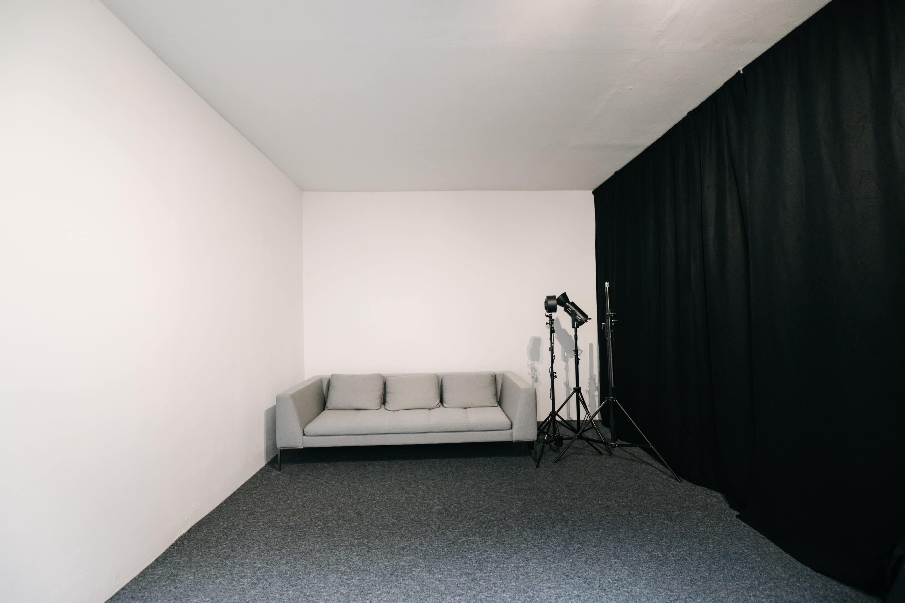 The image shows a simple room featuring a gray couch against a plain white wall, with a black curtain in one corner and photography equipment set up nearby.