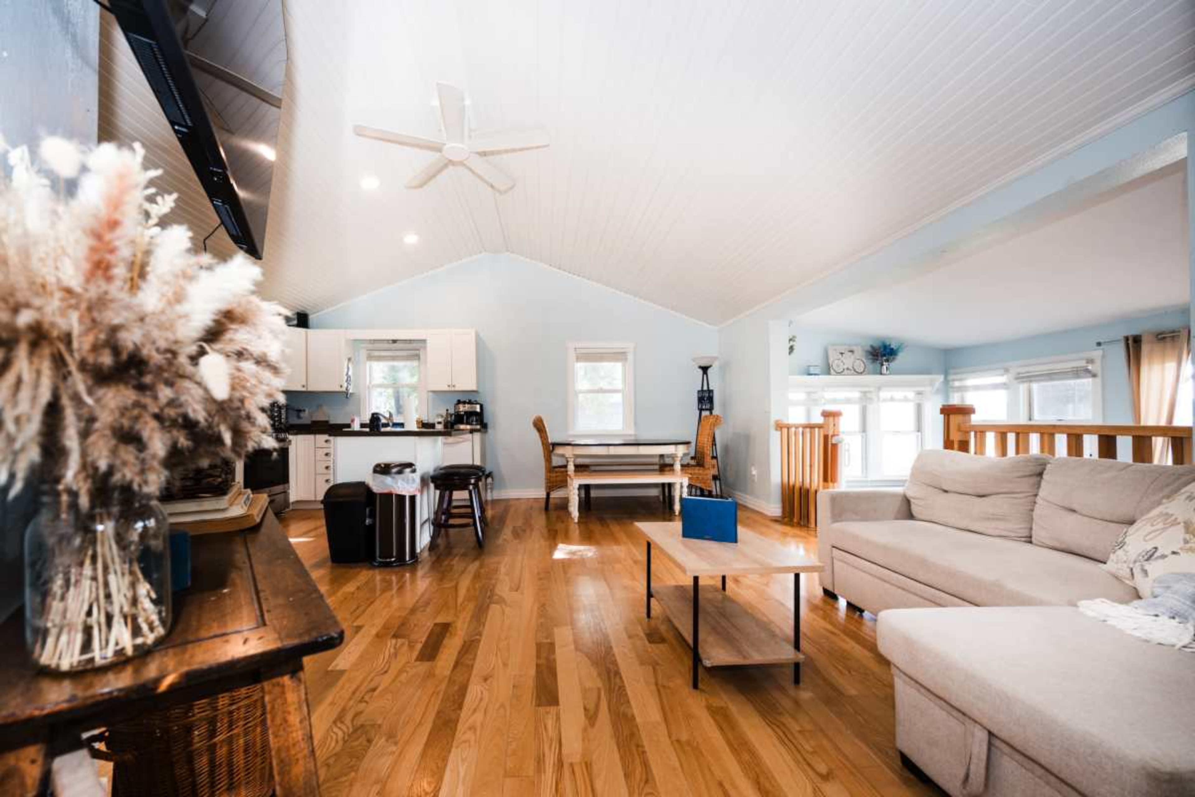 The image shows a spacious living area with a high ceiling, wooden flooring, and a light blue color scheme, featuring a sofa, coffee table, and kitchen area in the background.