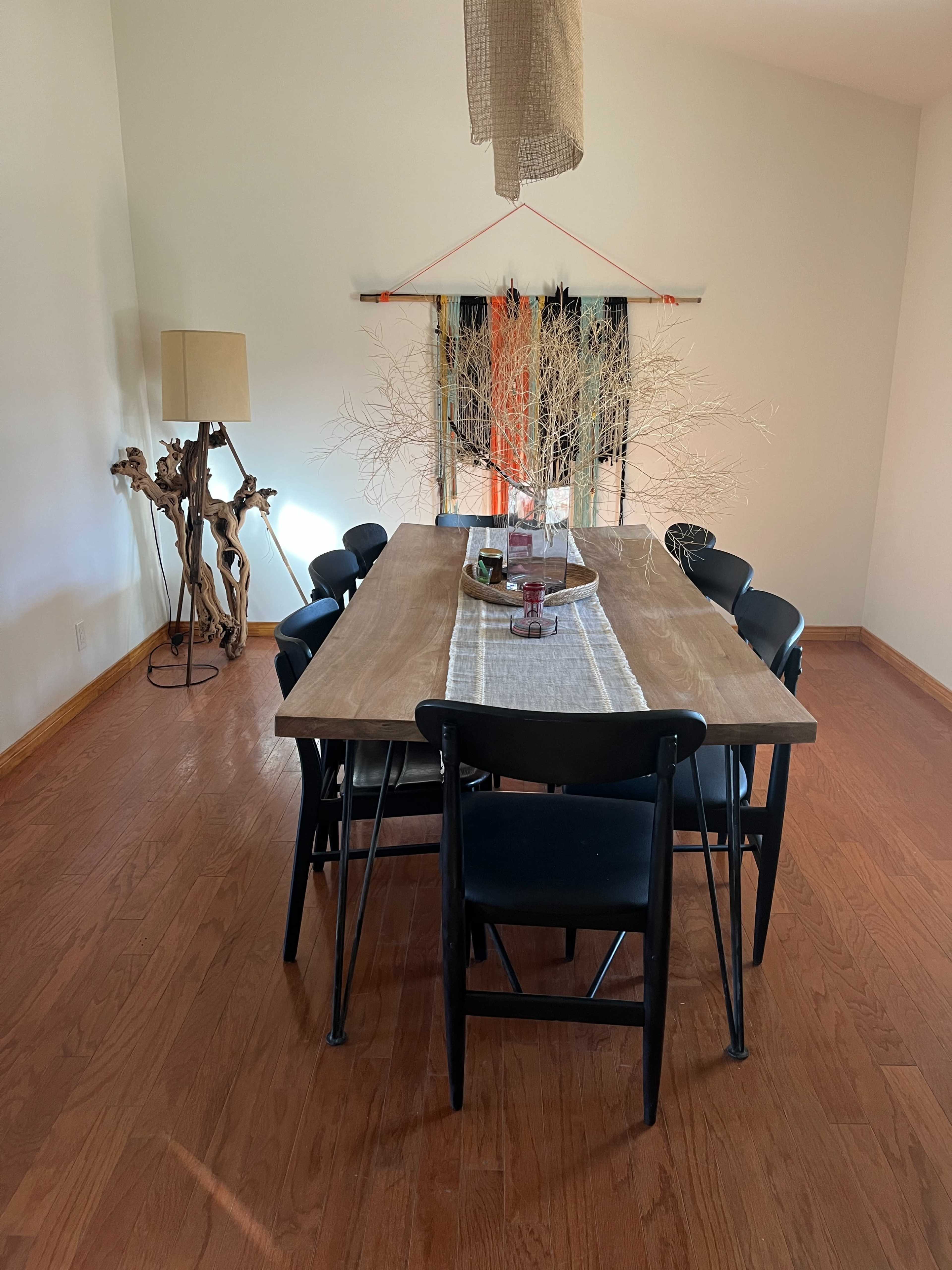A wooden dining table surrounded by black chairs is set in a bright room with a floor lamp and decorative branches in the center.