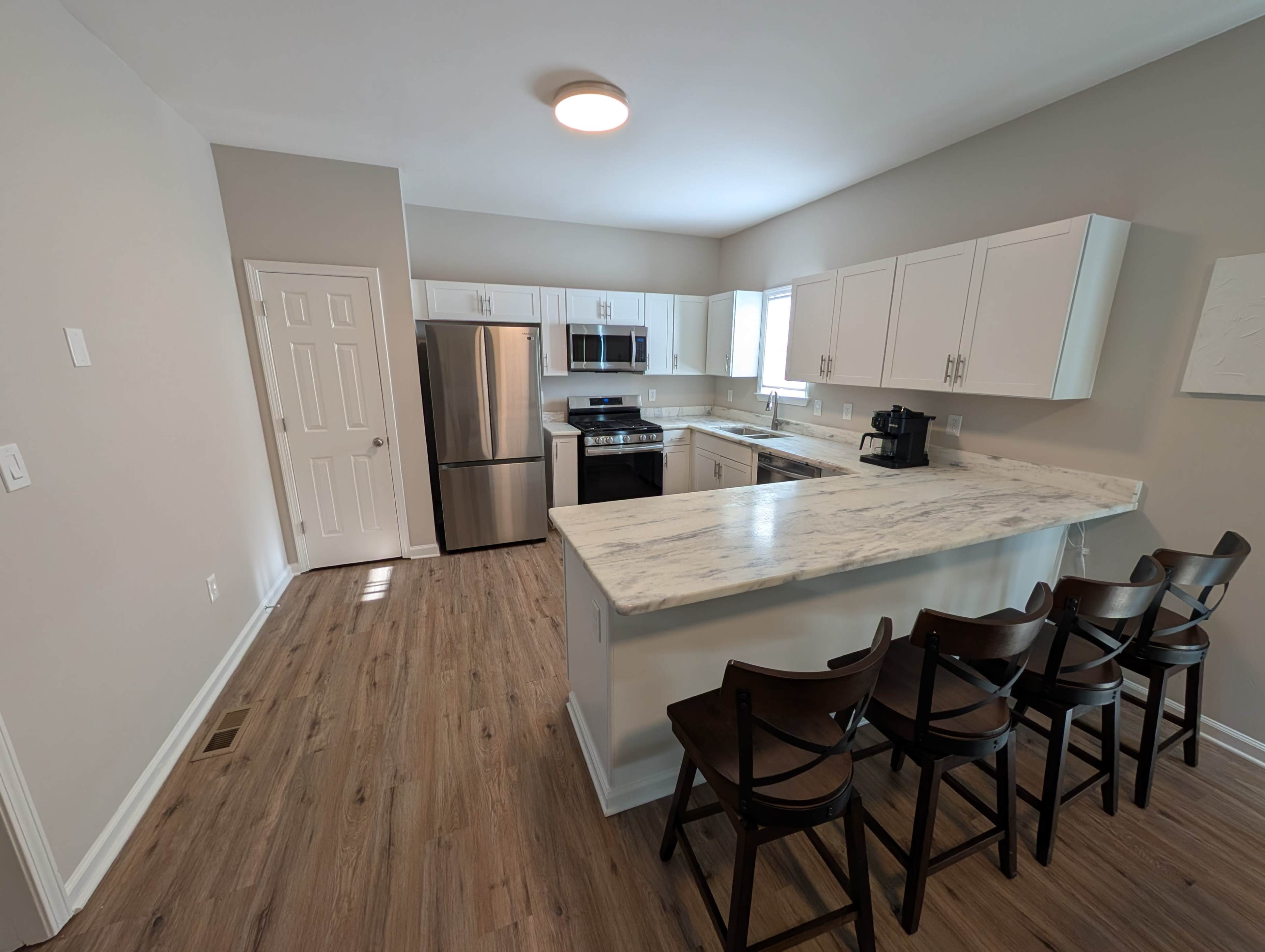 The image shows a modern kitchen with white cabinets, stainless steel appliances, and a marble countertop with bar seating.