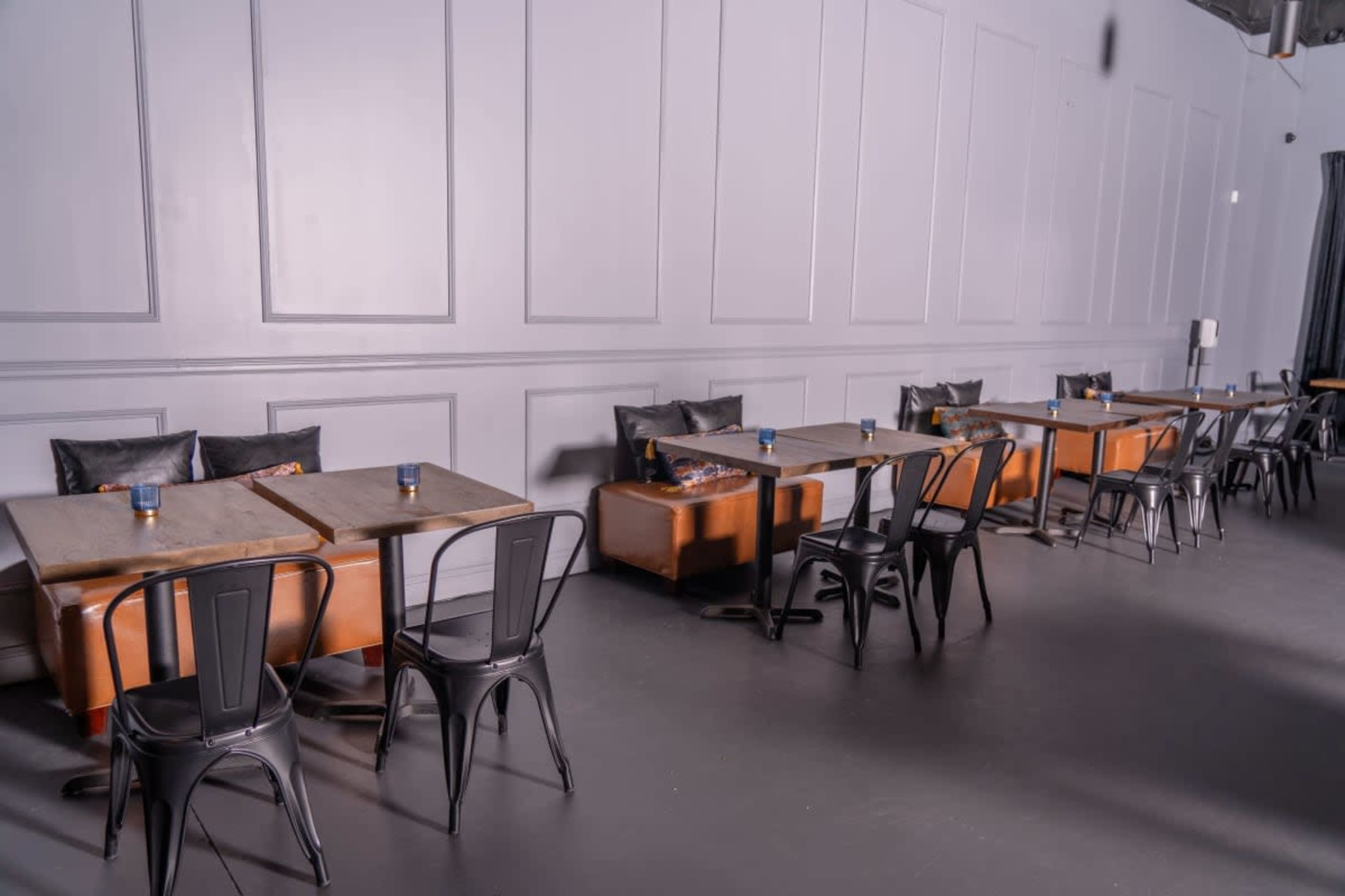 A minimalist indoor café space featuring wooden tables and black metal chairs arranged across a gray wall.