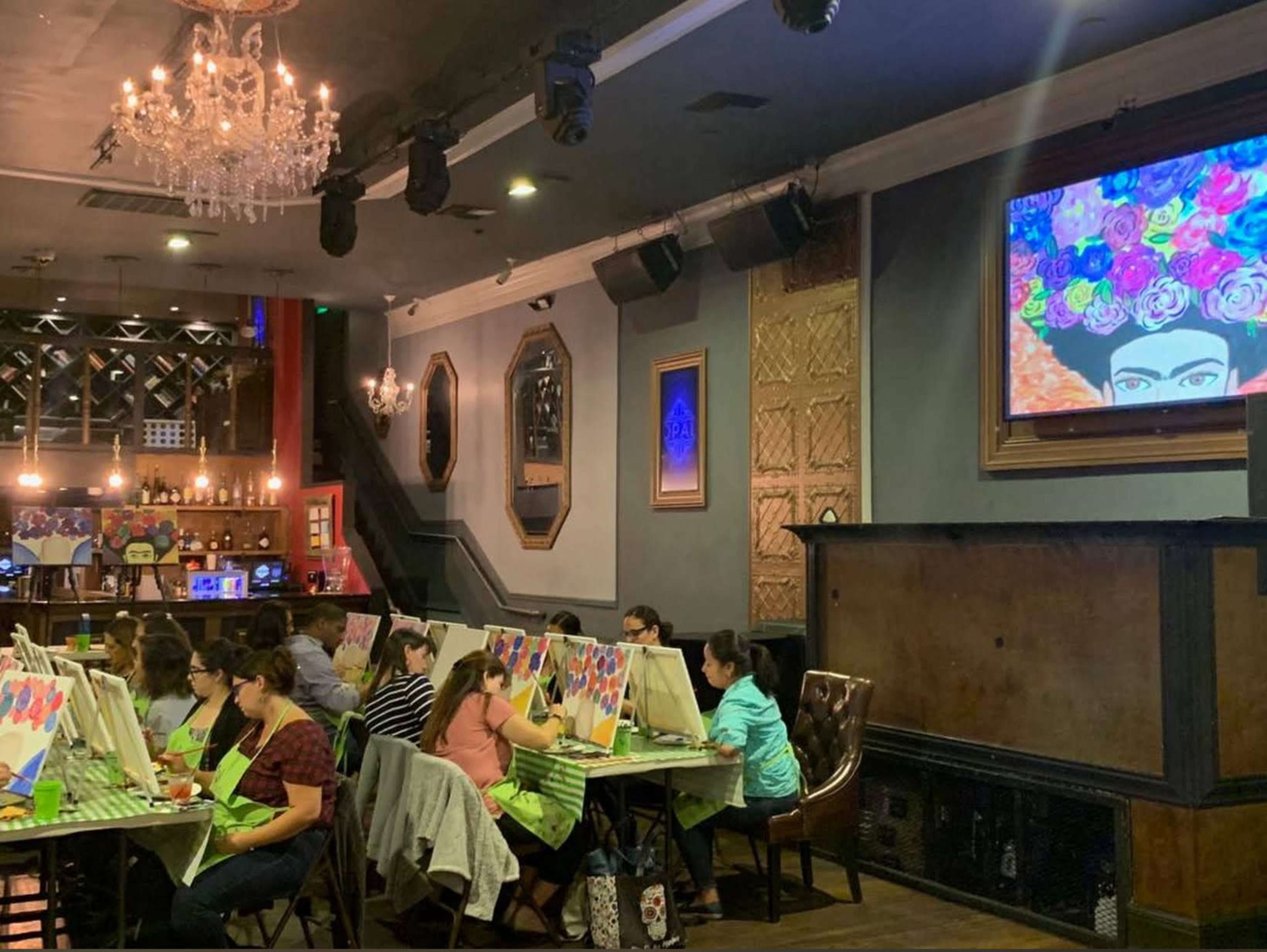 A group of people participates in a painting class in a dimly lit venue with a chandelier and artwork displayed on the wall.