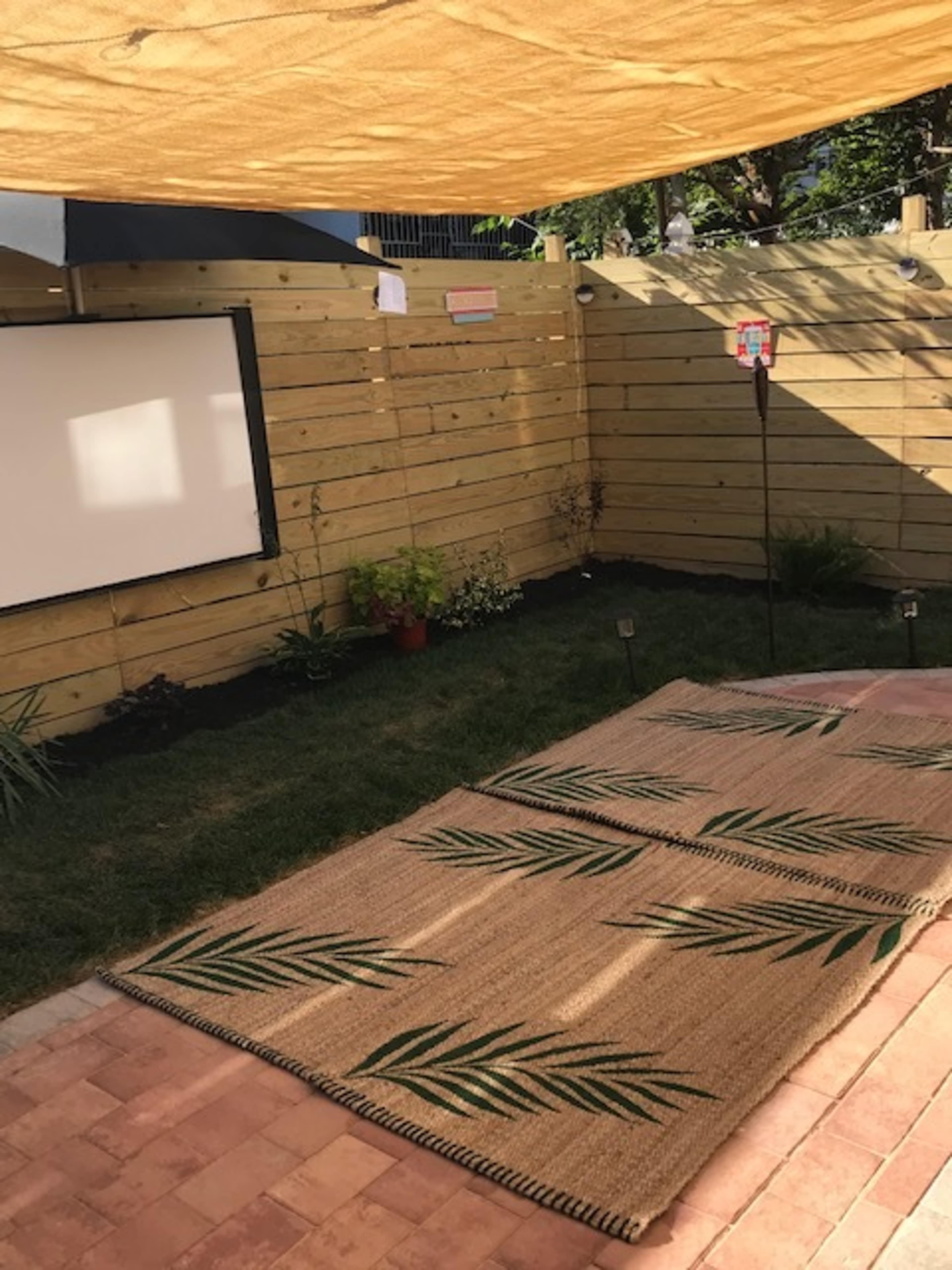 A small outdoor space with a grassy area, a decorative rug featuring leaf patterns, a blank screen mounted on a wooden fence, and potted plants.