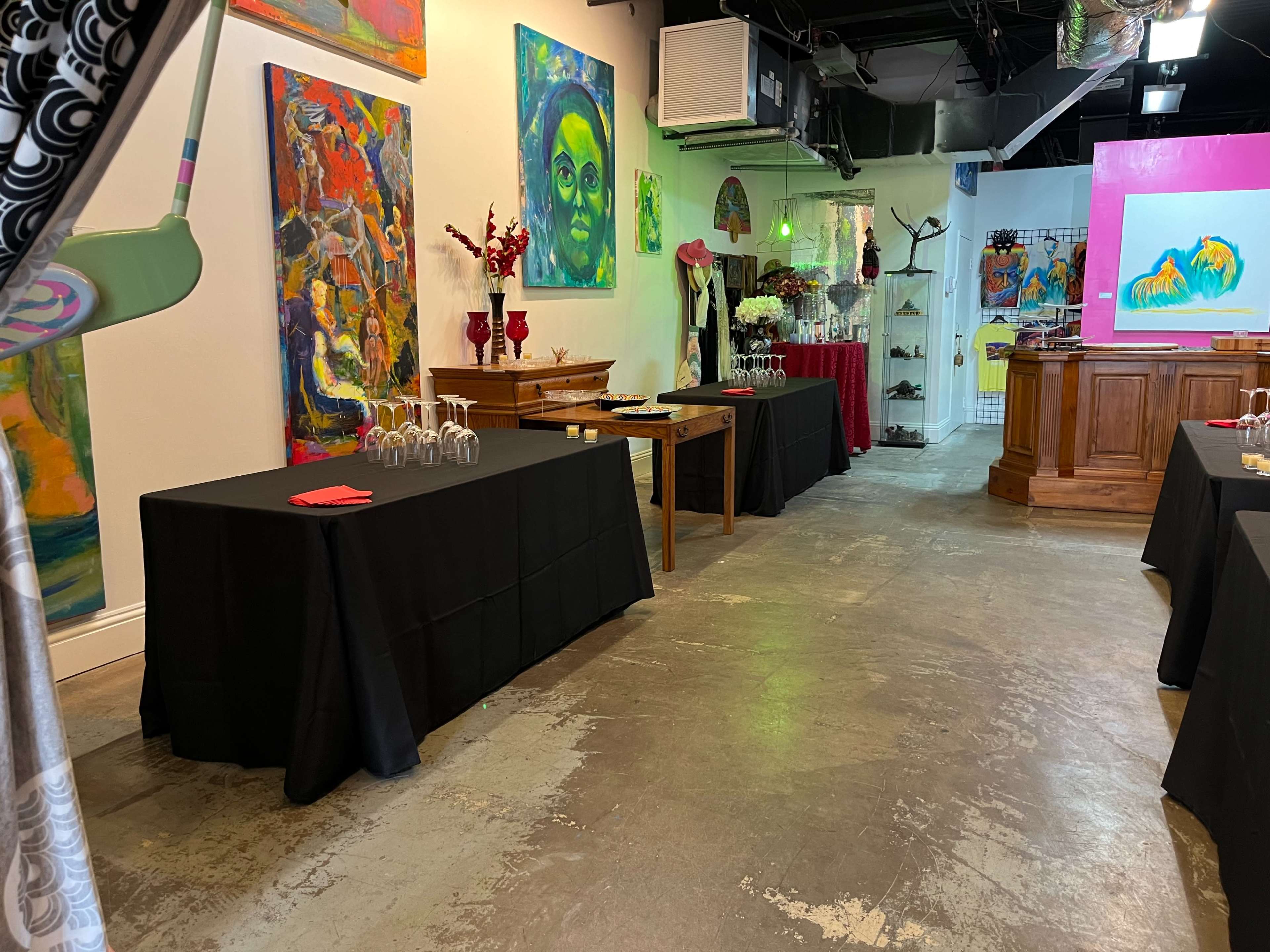An art gallery space with tables covered in black cloth, colorful paintings on the walls, and decorative items displayed on shelves.