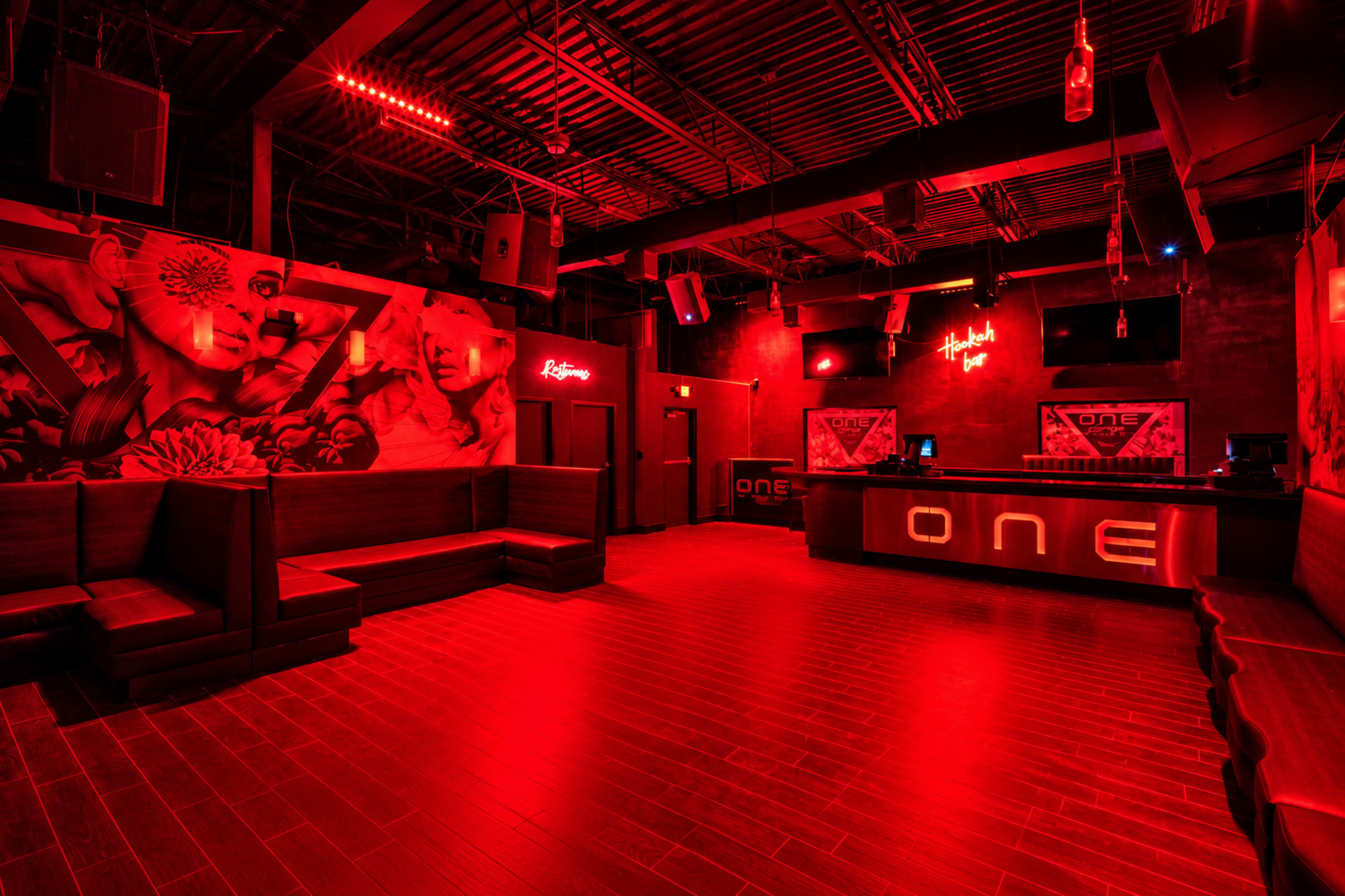 The image shows a dimly lit nightclub featuring red lighting, a bar area with the number "ONE" prominently displayed, and mural artwork on the walls.