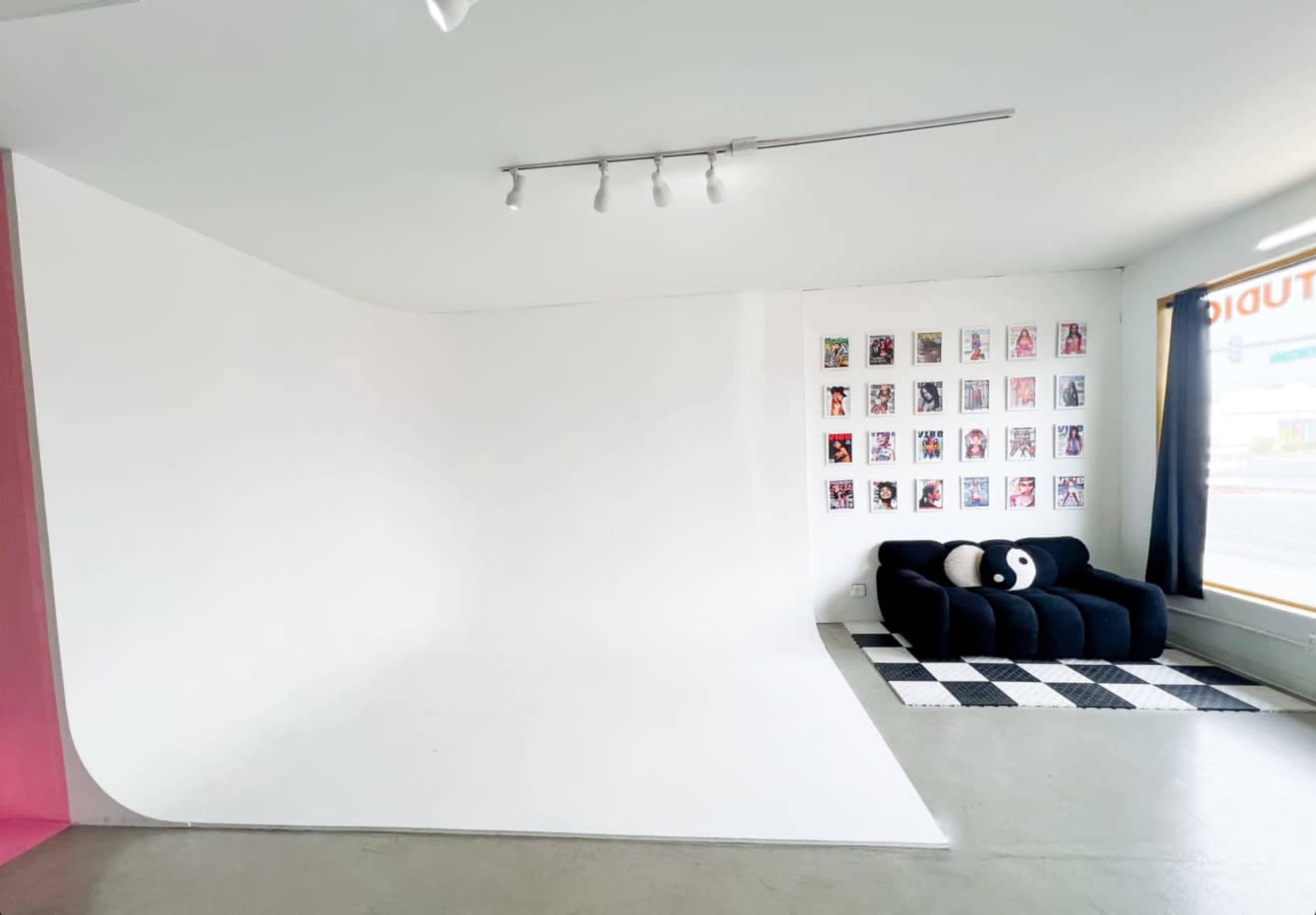 The image shows a bright, minimalist studio space with a curved white backdrop, a black couch, and a gallery wall of framed photos.