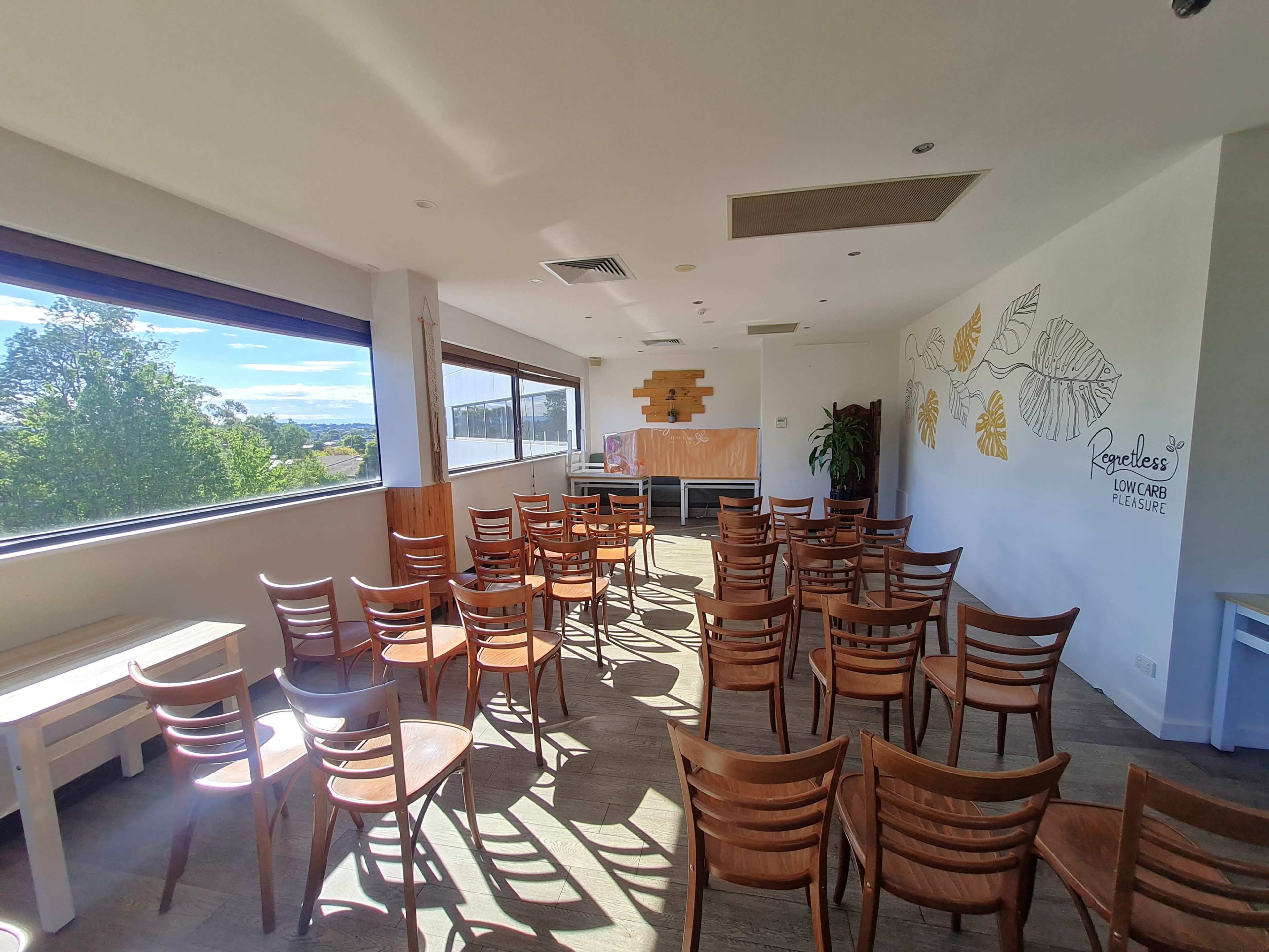 The image shows a spacious room with several rows of wooden chairs arranged facing a blank wall and large windows that allow natural light to enter.