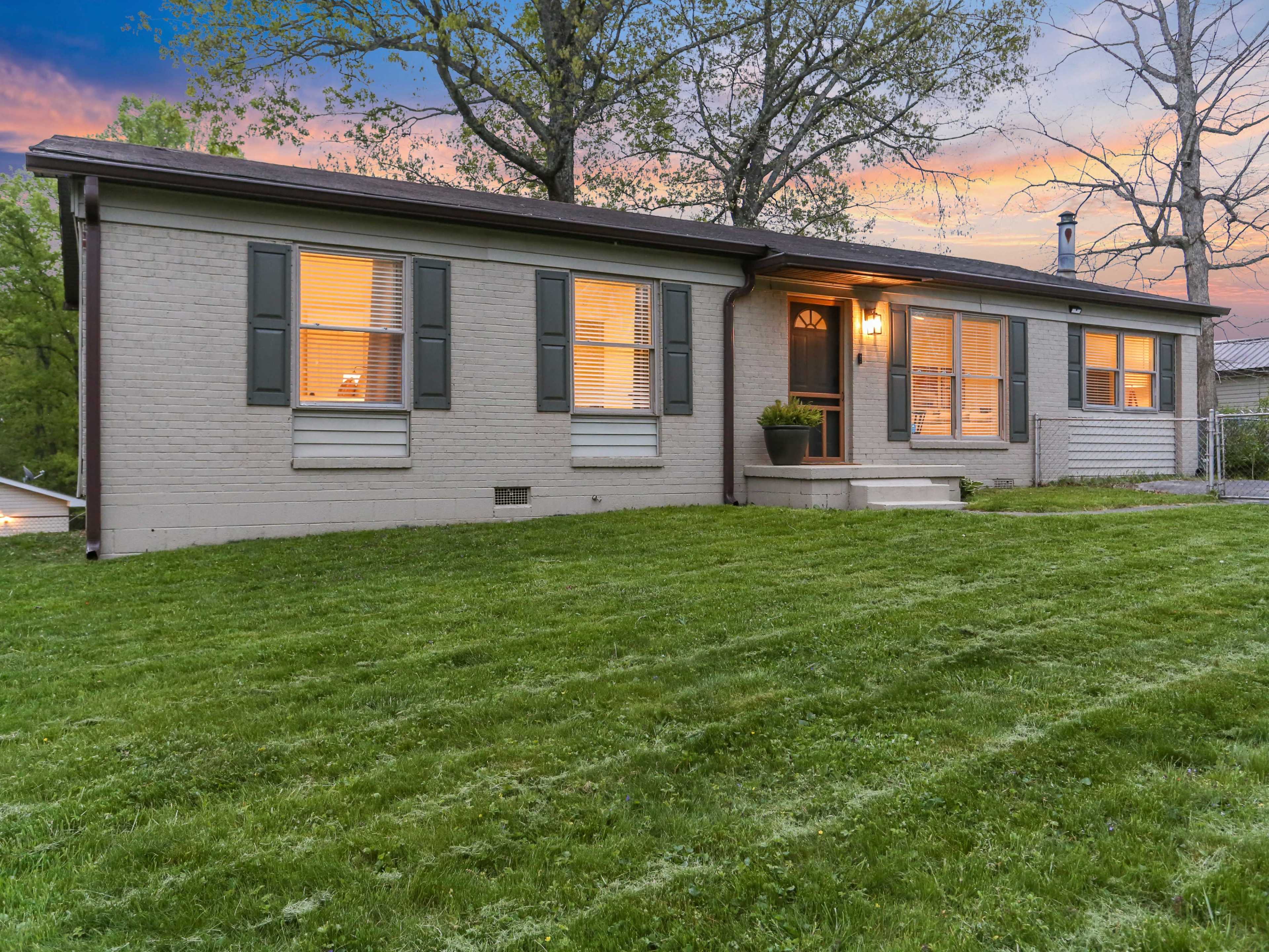 The image shows a single-story, brick house with green shutters and a well-maintained lawn, illuminated by evening light.