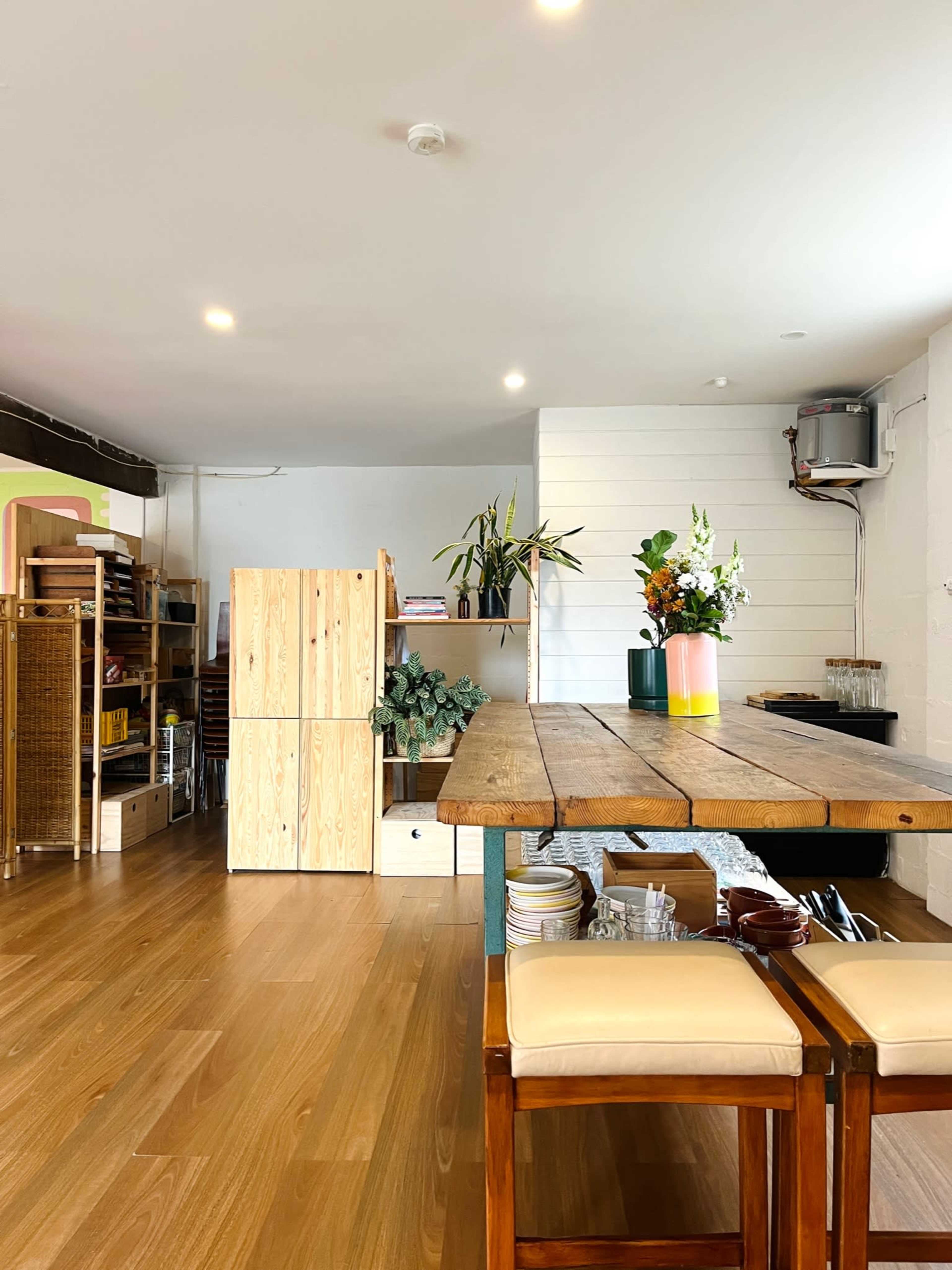 The image shows a minimalist interior of a cafe or workspace with a wooden table, seating, and shelves lined with plants and various items.