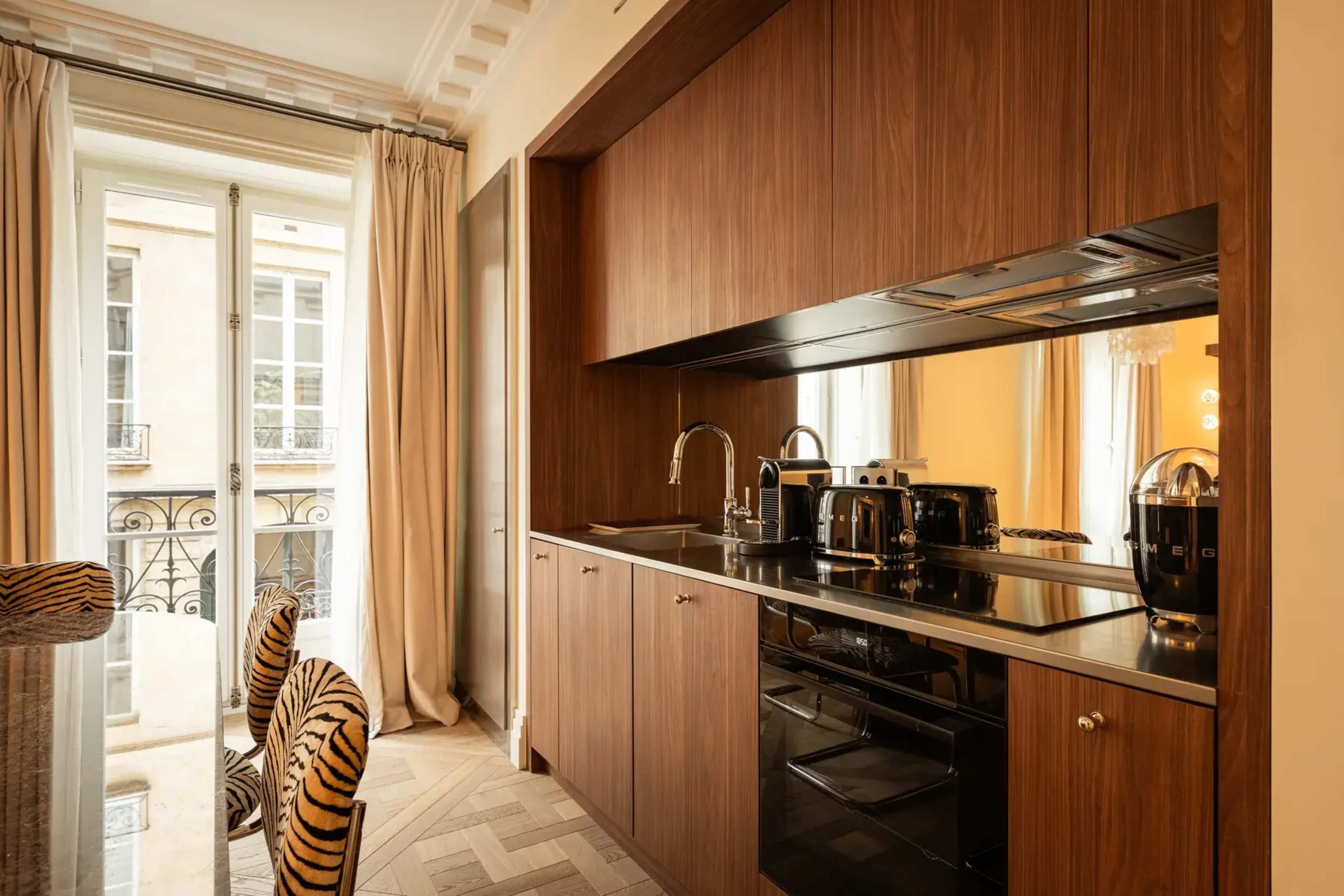 The image shows a modern kitchen area featuring wooden cabinetry, a black countertop with built-in appliances, and large windows with sheer curtains.