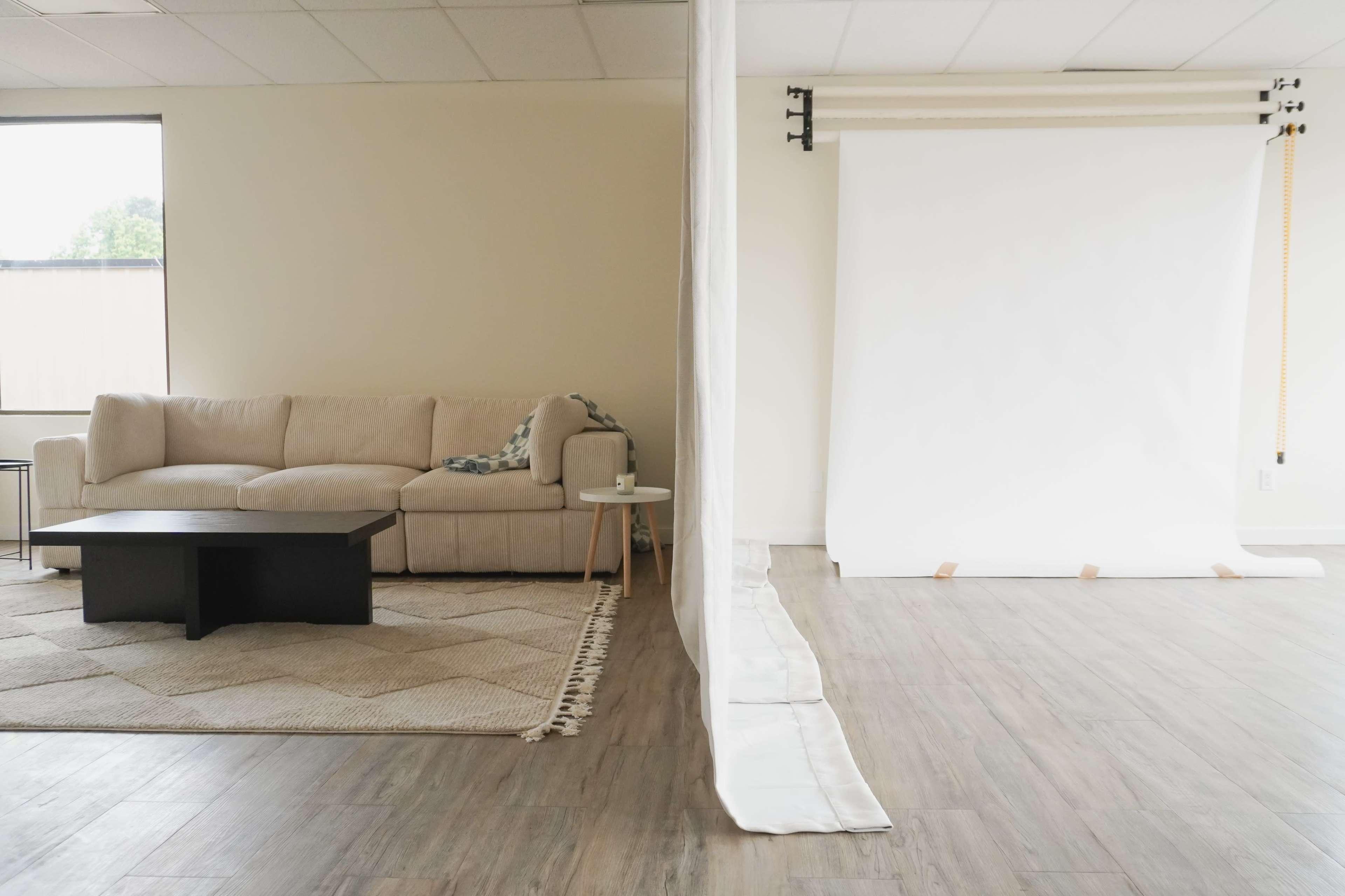 A spacious room divided by a white curtain, featuring a beige sofa, a black coffee table, and a white backdrop set up on one side.