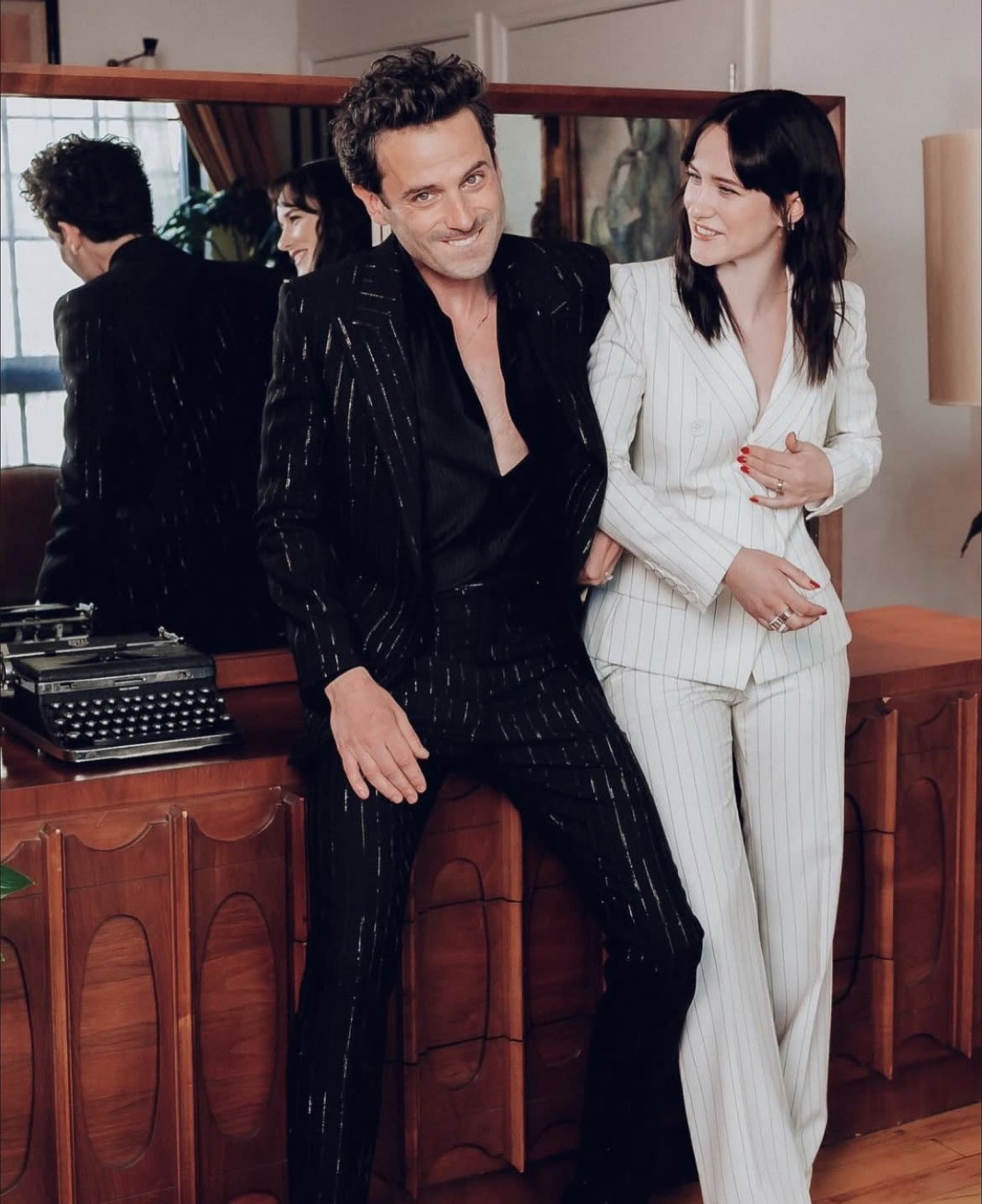 Two individuals stand together in a stylish interior, one wearing a black suit with pinstripes and the other in a white pinstripe suit.
