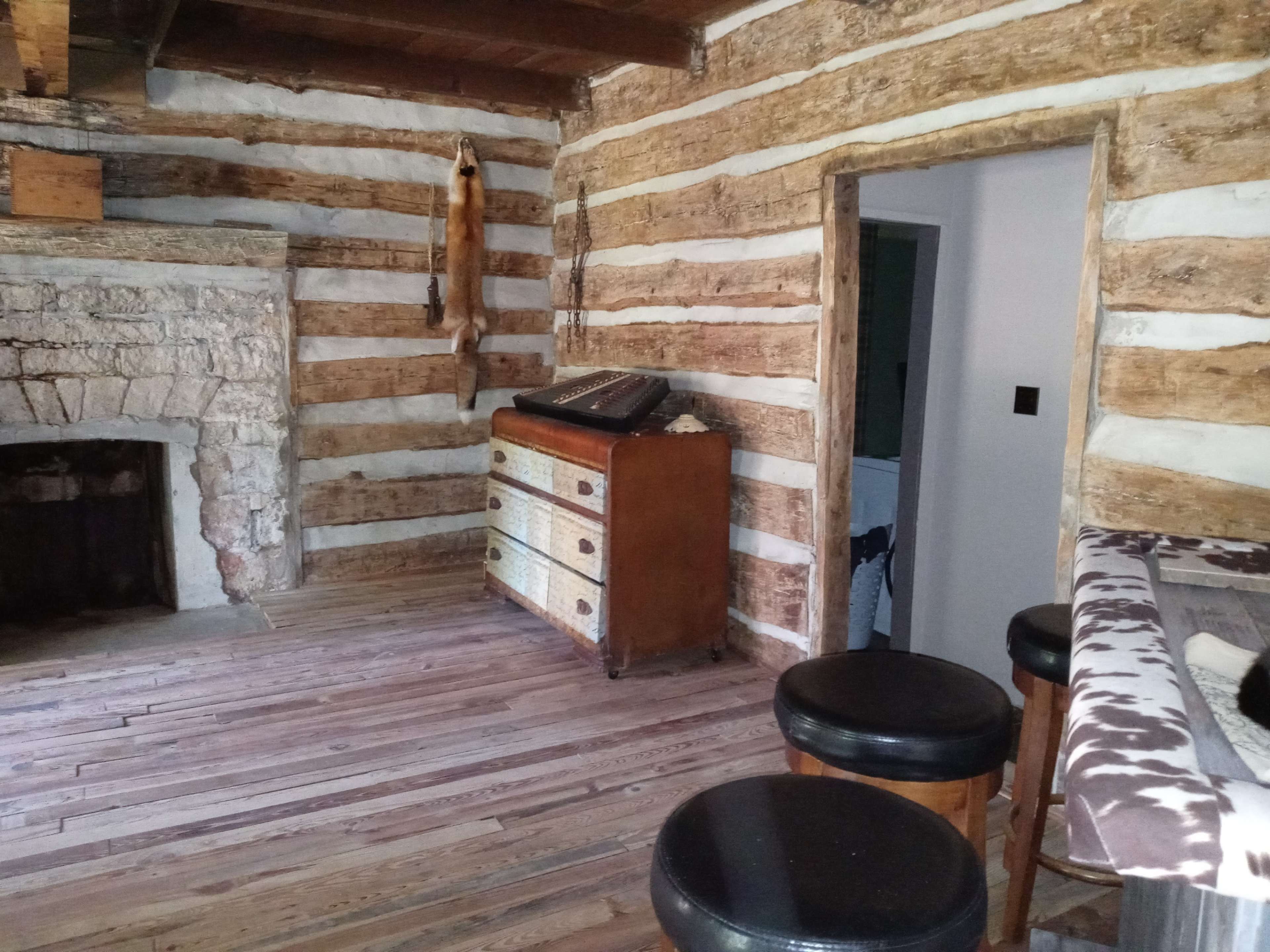The interior of a rustic cabin features log walls, a stone fireplace, a wooden dresser, and a small bar with stools.