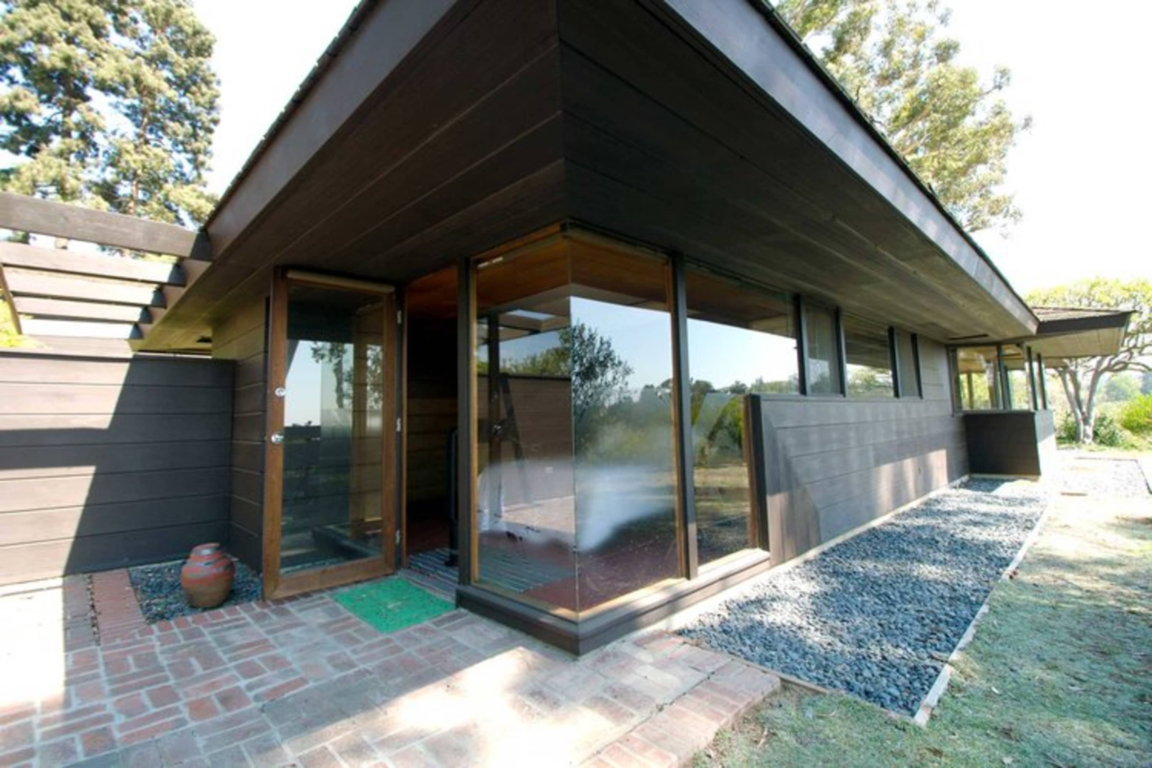 The image shows a modern, single-story house with large glass windows and a combination of wood and stone materials in its exterior design.