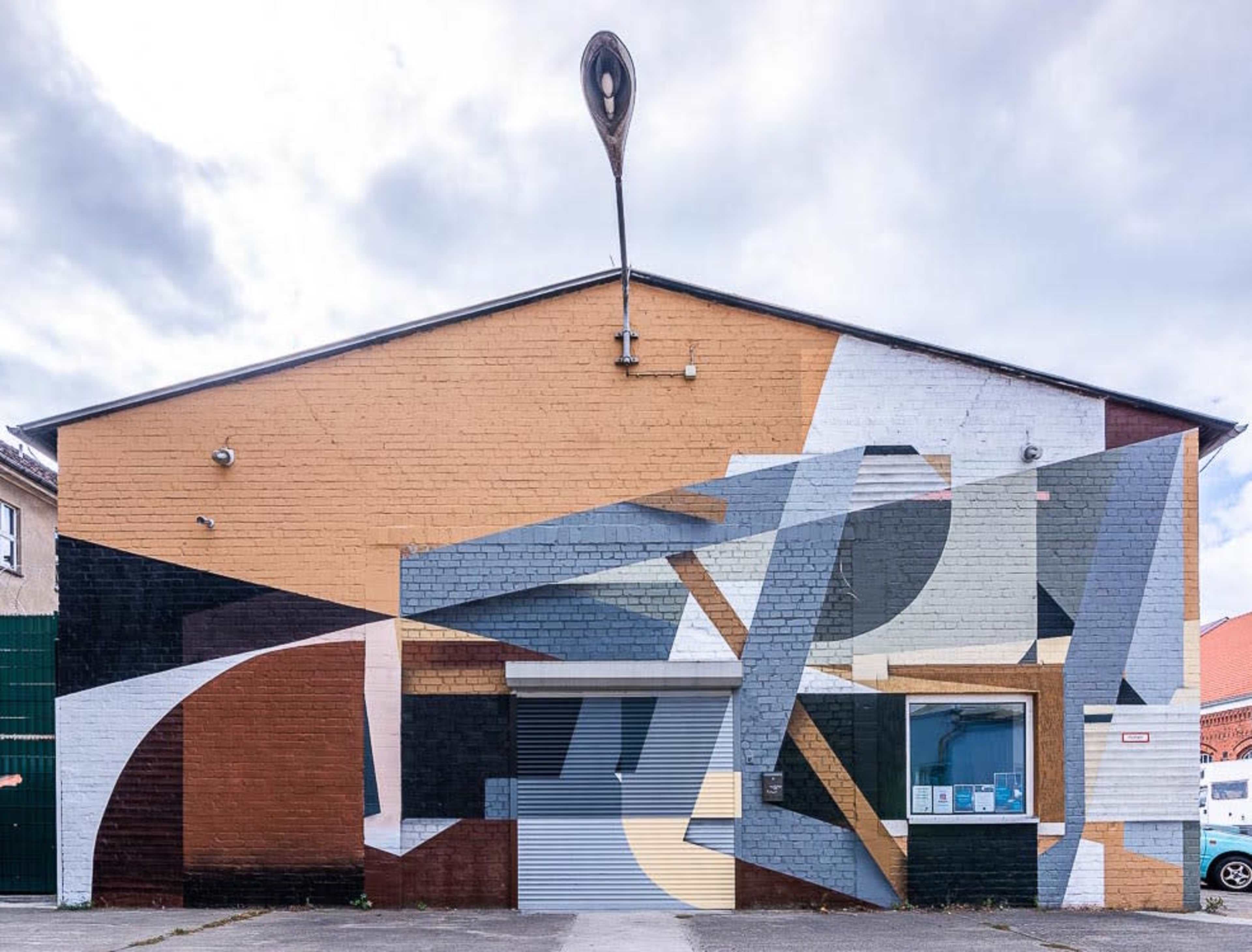 A large building with a geometric mural in orange, black, and gray on its facade, featuring a prominent spoon-shaped structure at the top.