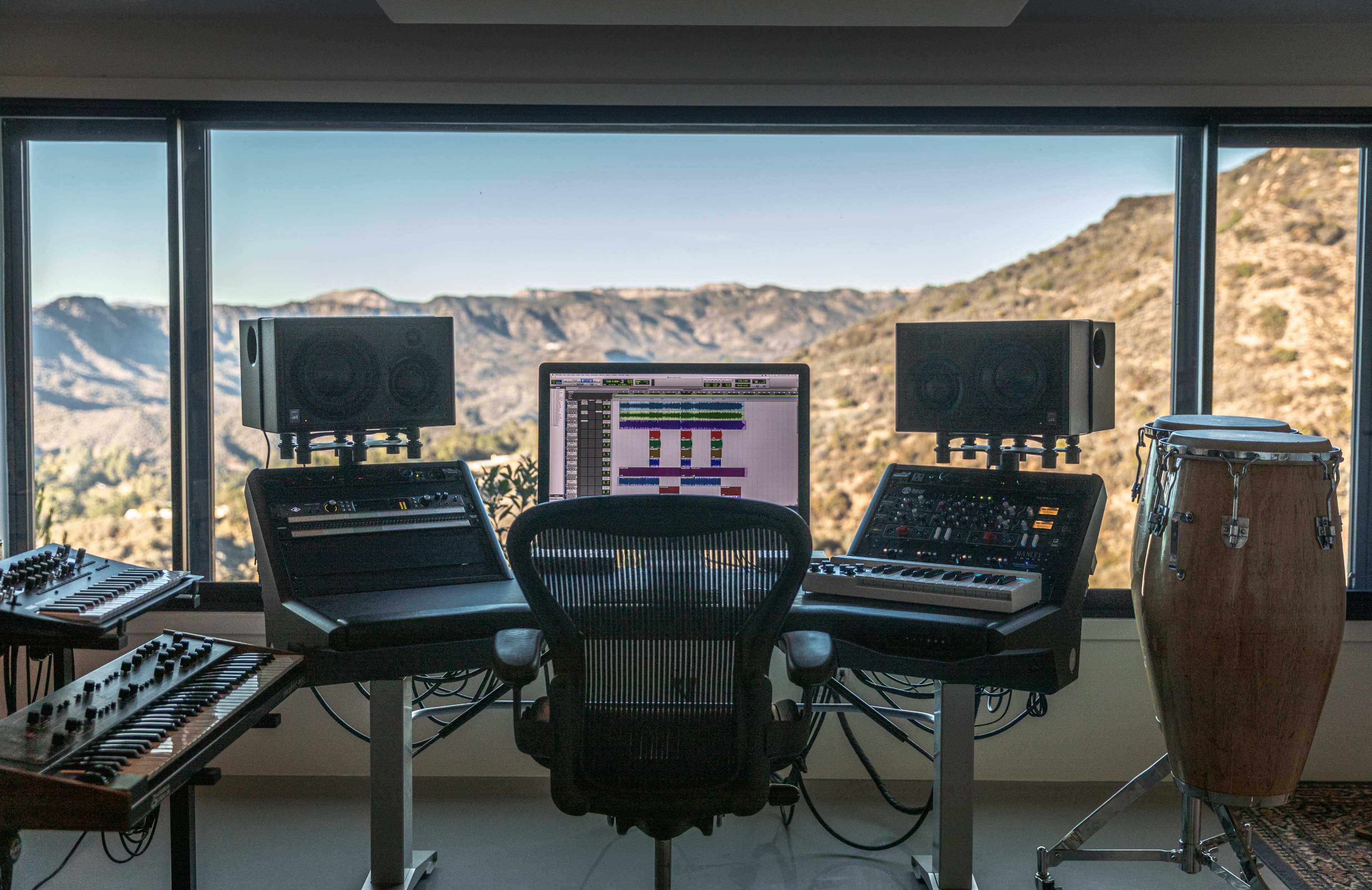 A modern music studio is set up with mixing equipment and instruments, facing large windows that showcase a mountain landscape.