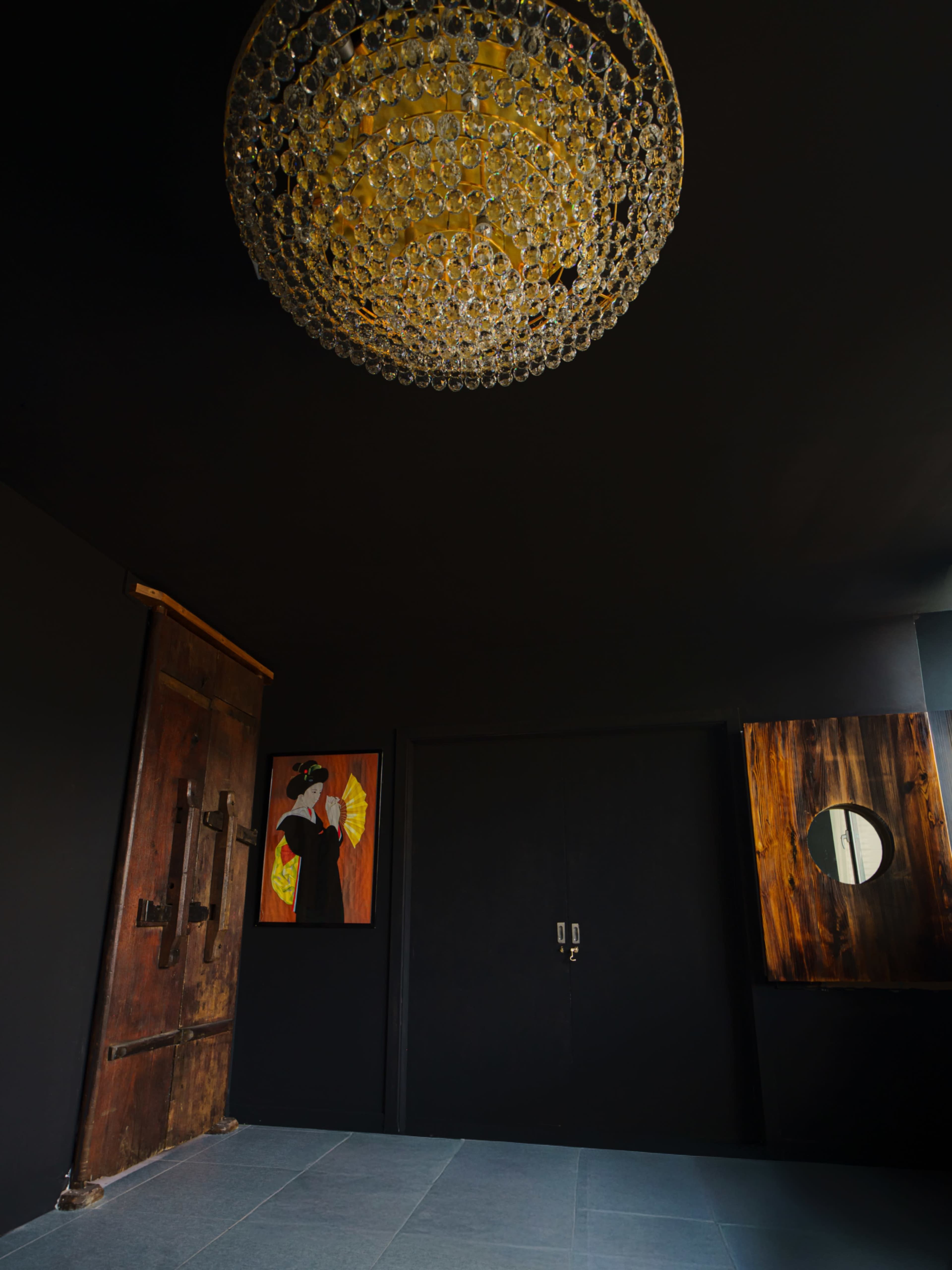 The image shows a dimly lit room with a large chandelier, a wooden door on the left, a black wall with a painting, and a round mirror on the right.