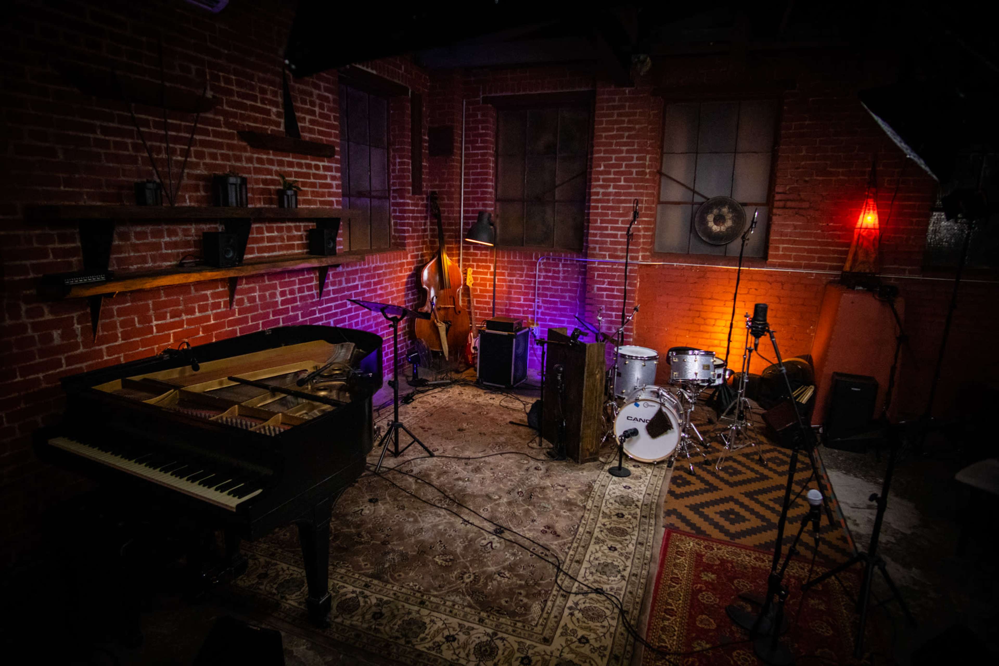 A dimly lit music studio featuring a piano, a double bass, drum set, and various sound equipment against a backdrop of exposed red brick walls.