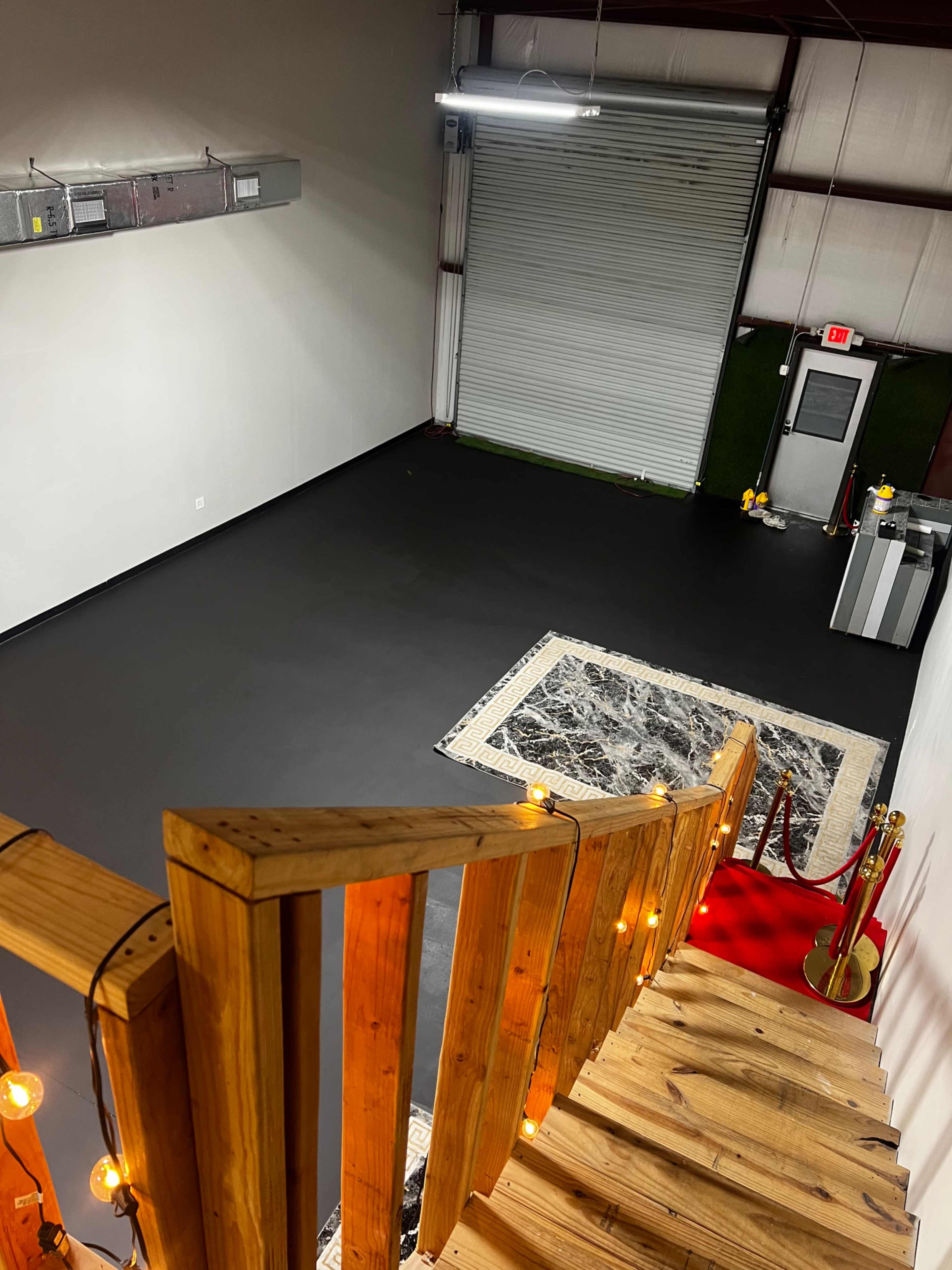 The image shows a spacious room with a polished dark floor, a large garage door, a decorative rug, and wooden stairs with string lights leading down from an upper level.