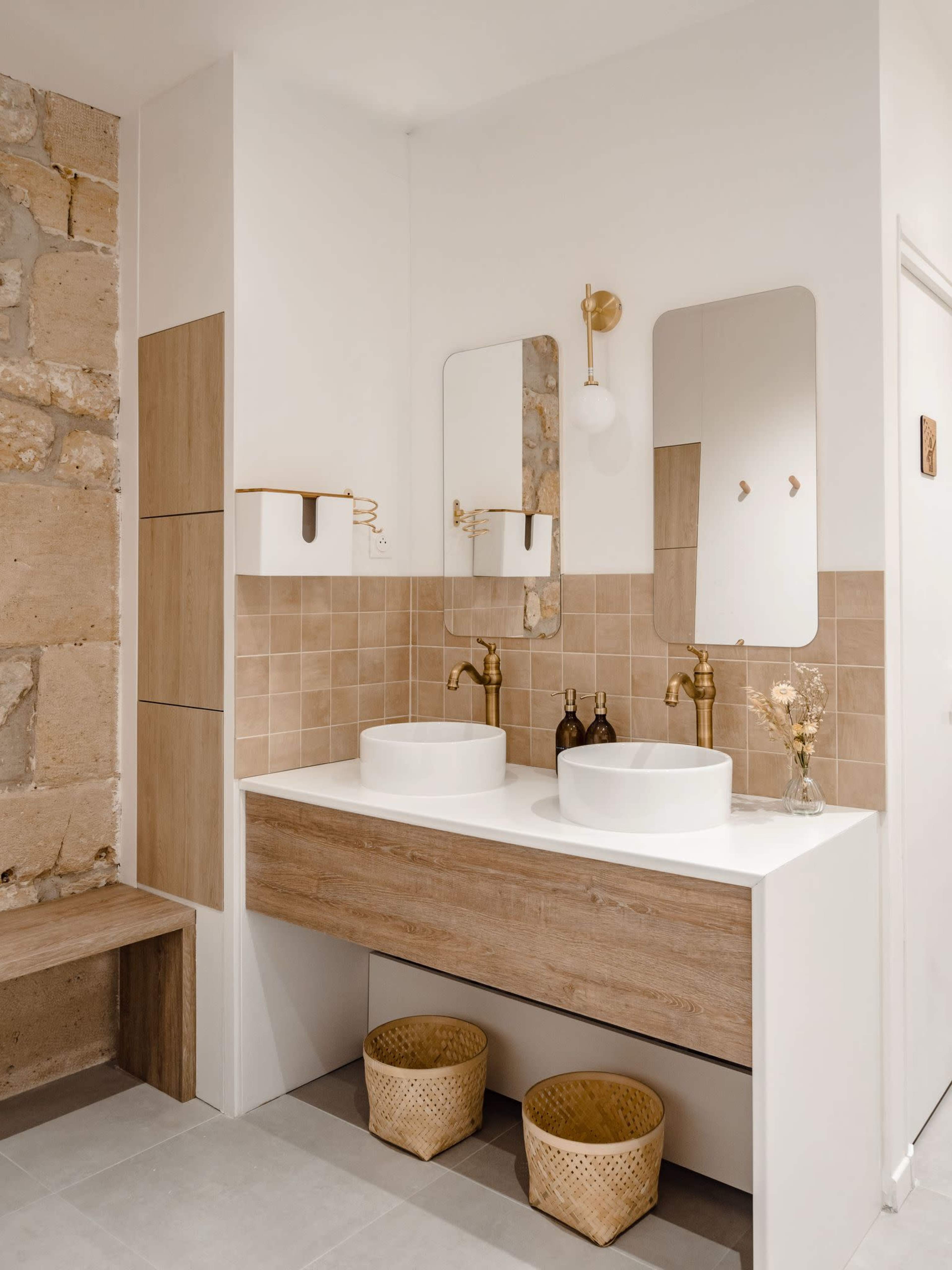 The image shows a modern bathroom featuring a double sink with round basins, gold faucets, two mirrors, and tiled walls in neutral tones.
