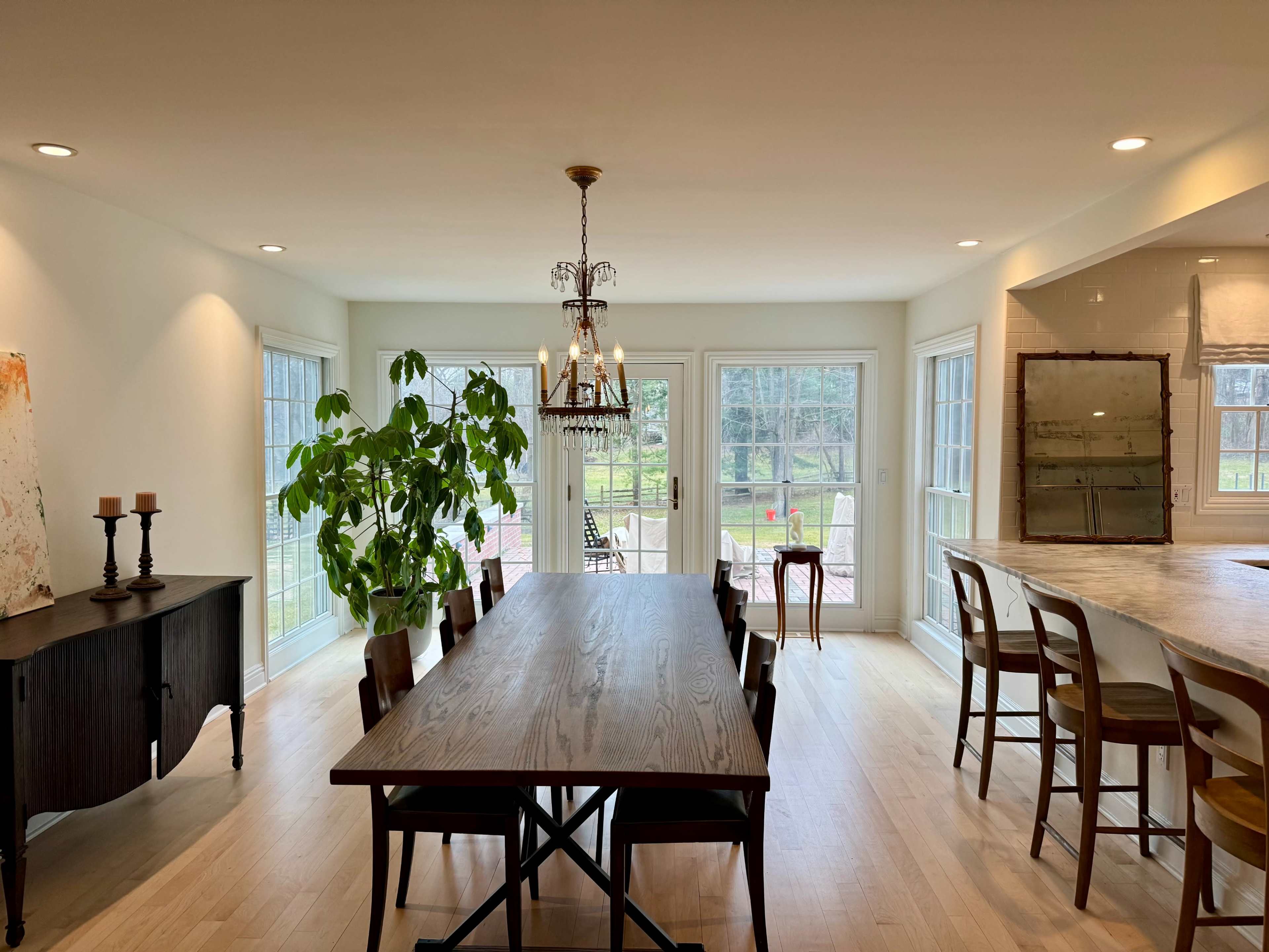 The image shows a bright dining area with a large wooden table, green plants, and large windows overlooking an outdoor space.