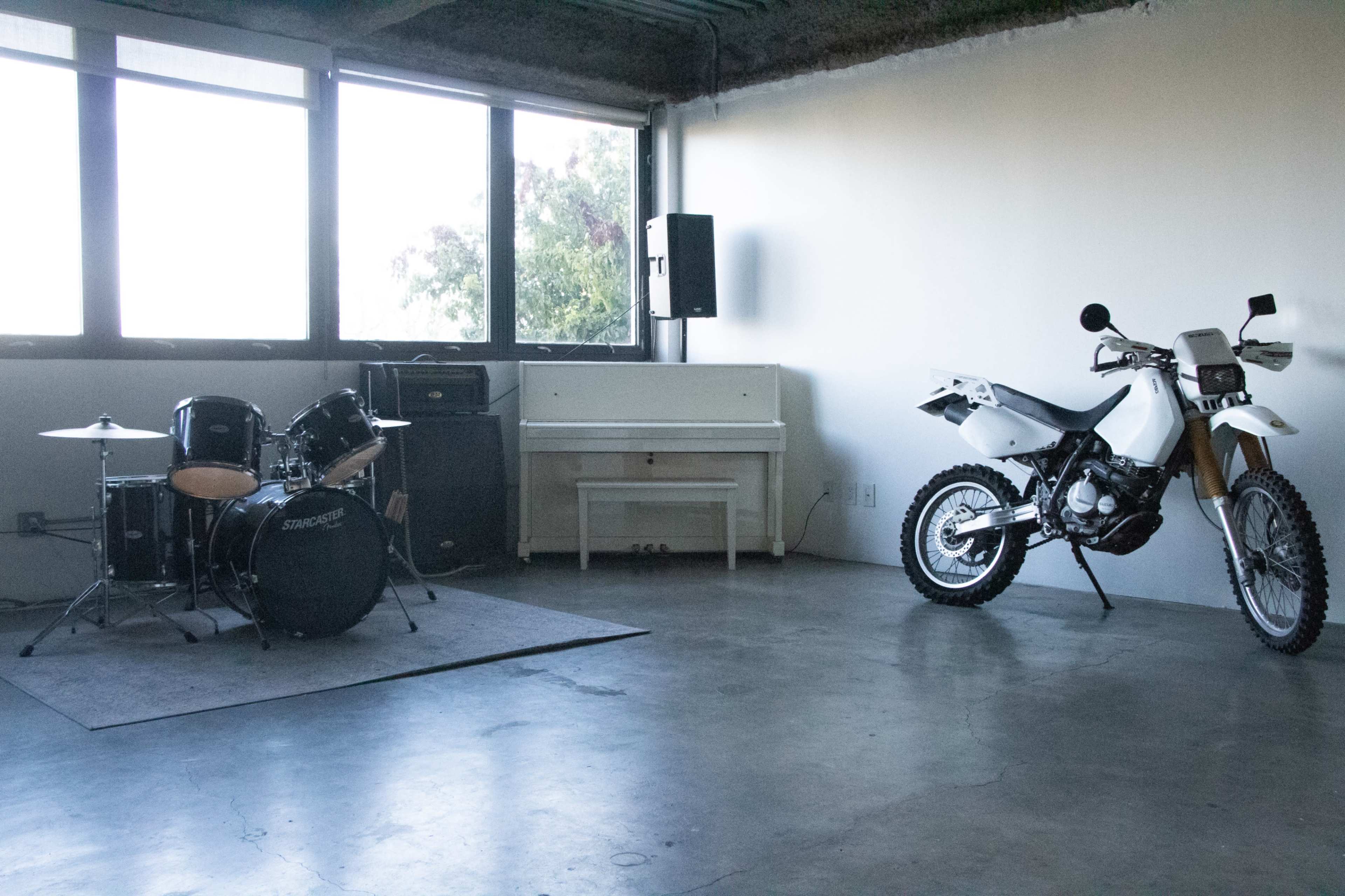 A room with a drum set, a motorcycle, and a white piano against bare walls and large windows.