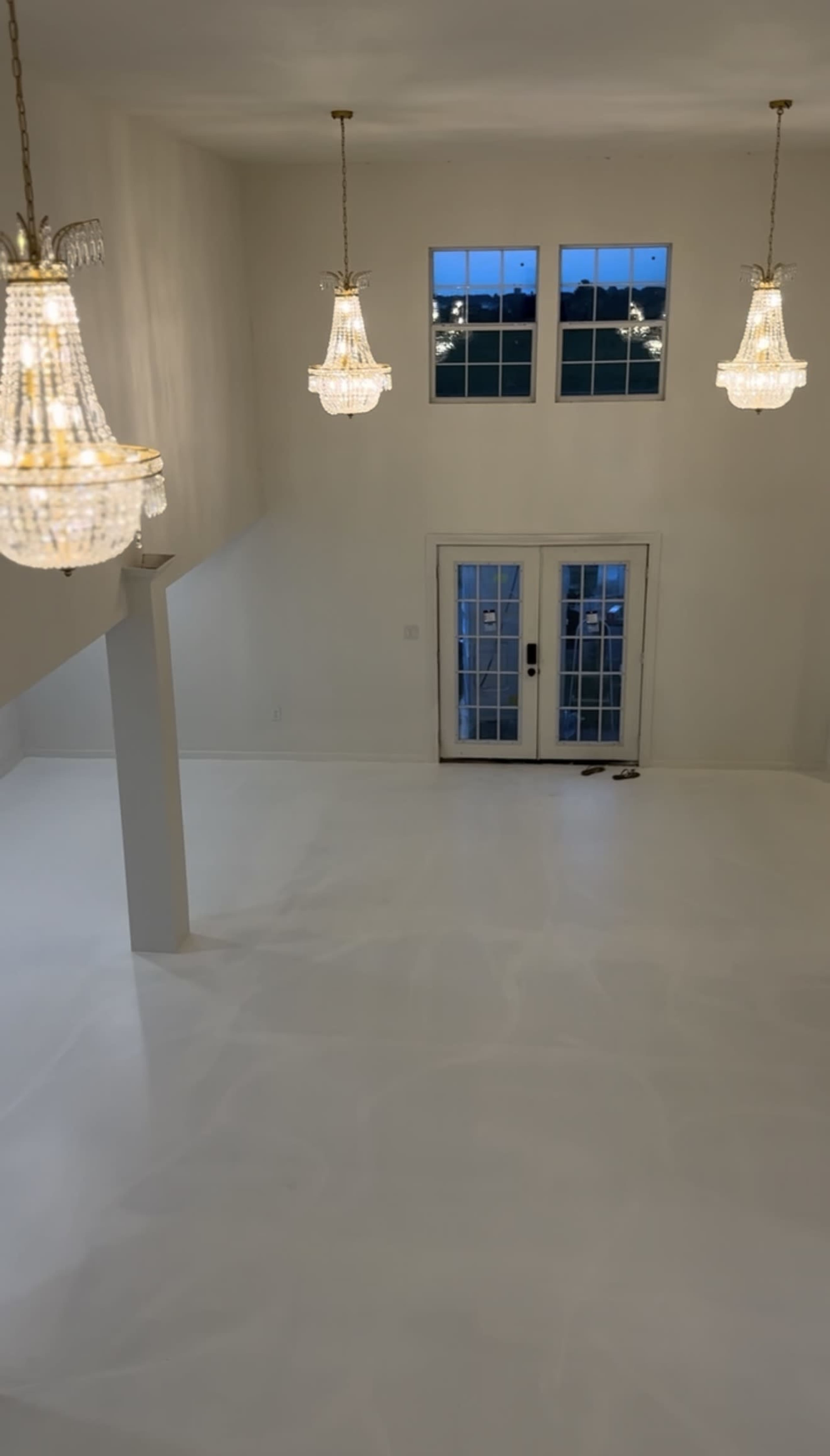 The image shows a spacious, minimally decorated interior room with white walls and a glossy white floor, illuminated by several ornate chandeliers.