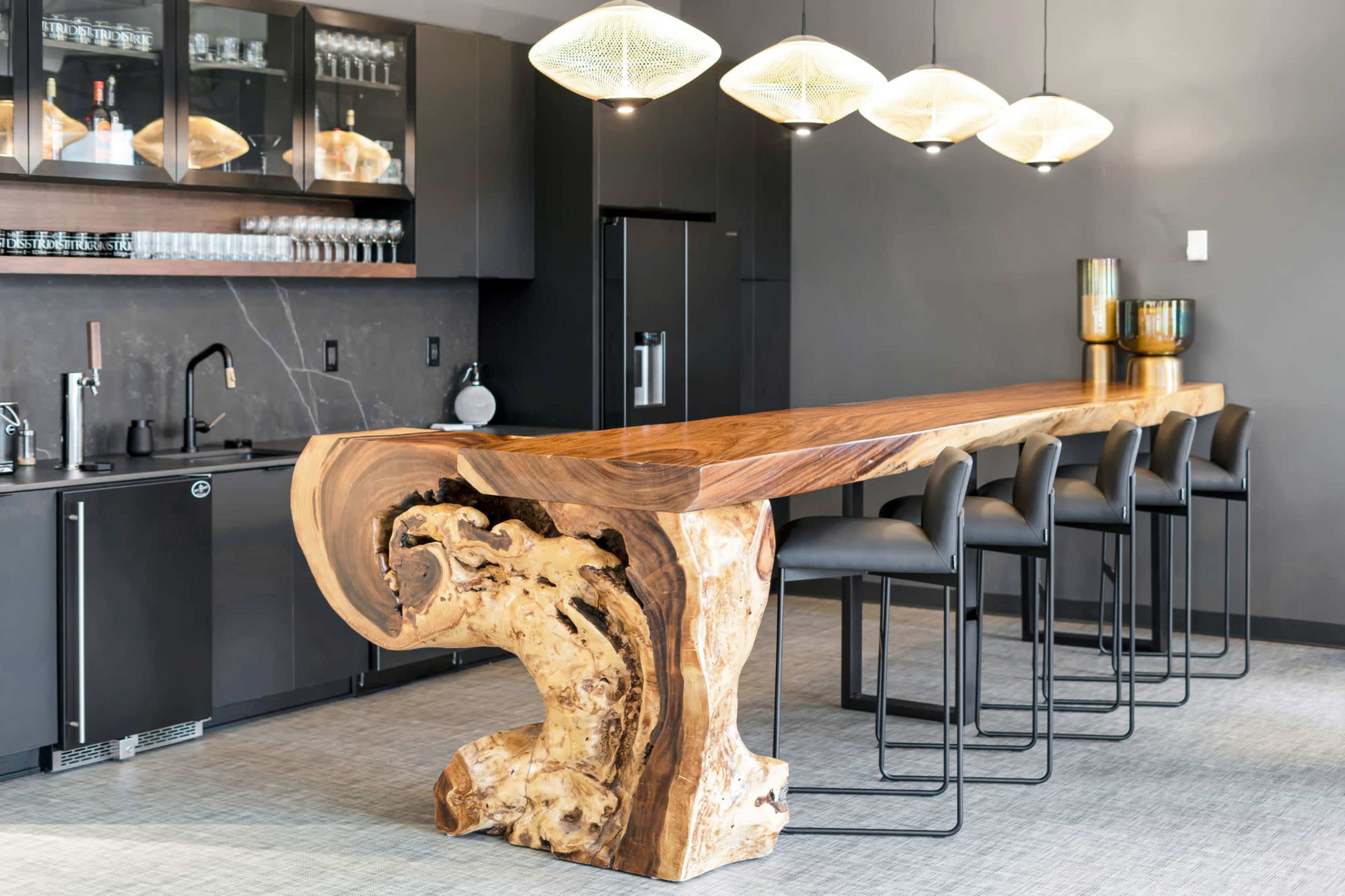 The image shows a modern kitchen bar featuring a large wooden countertop supported by a sculptural base, with four black stools positioned along one side.